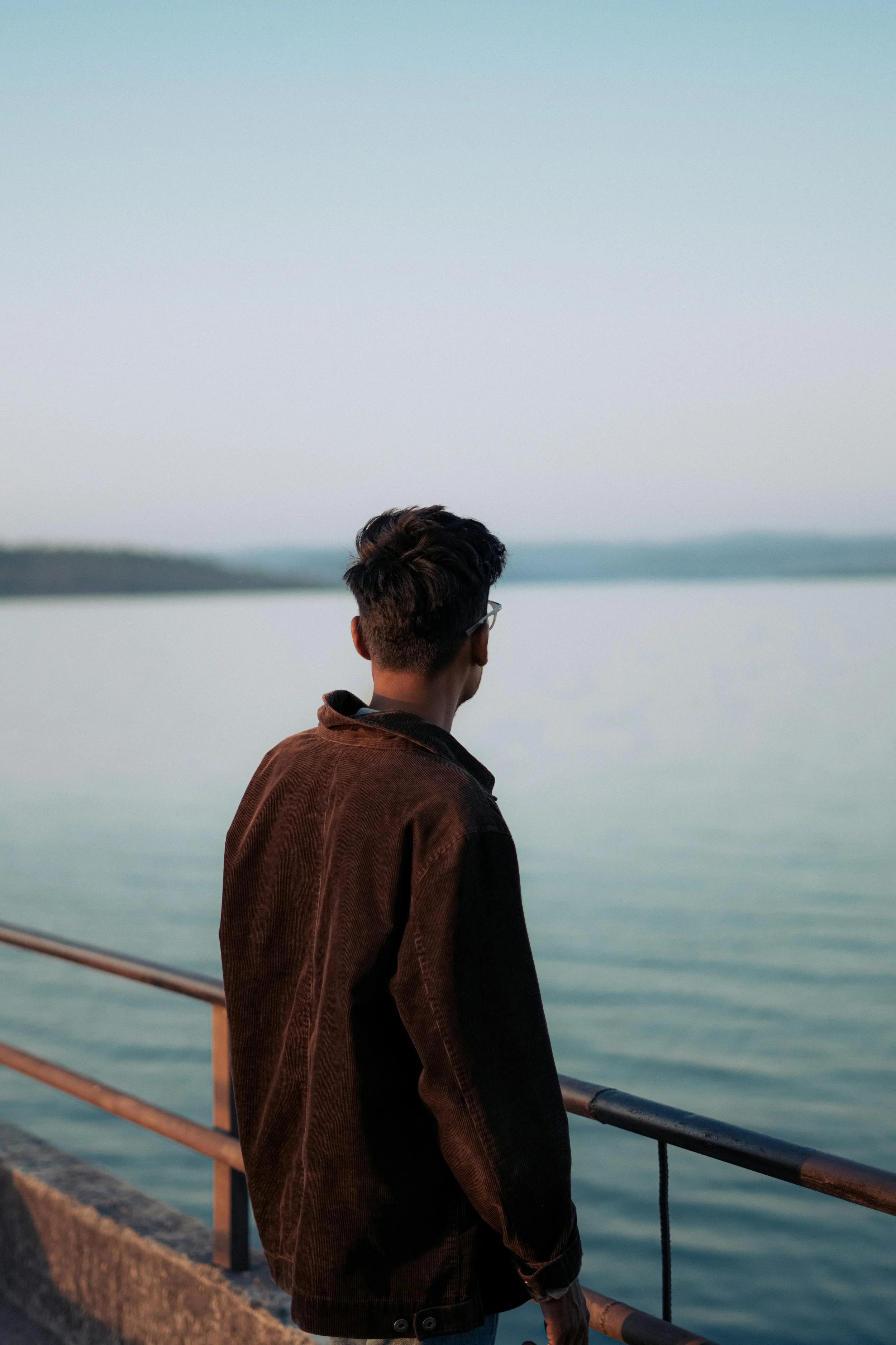 A young man with dark hair wearing glasses and a brown jacket stands on a pier, looking out over a calm body of water during sunset.