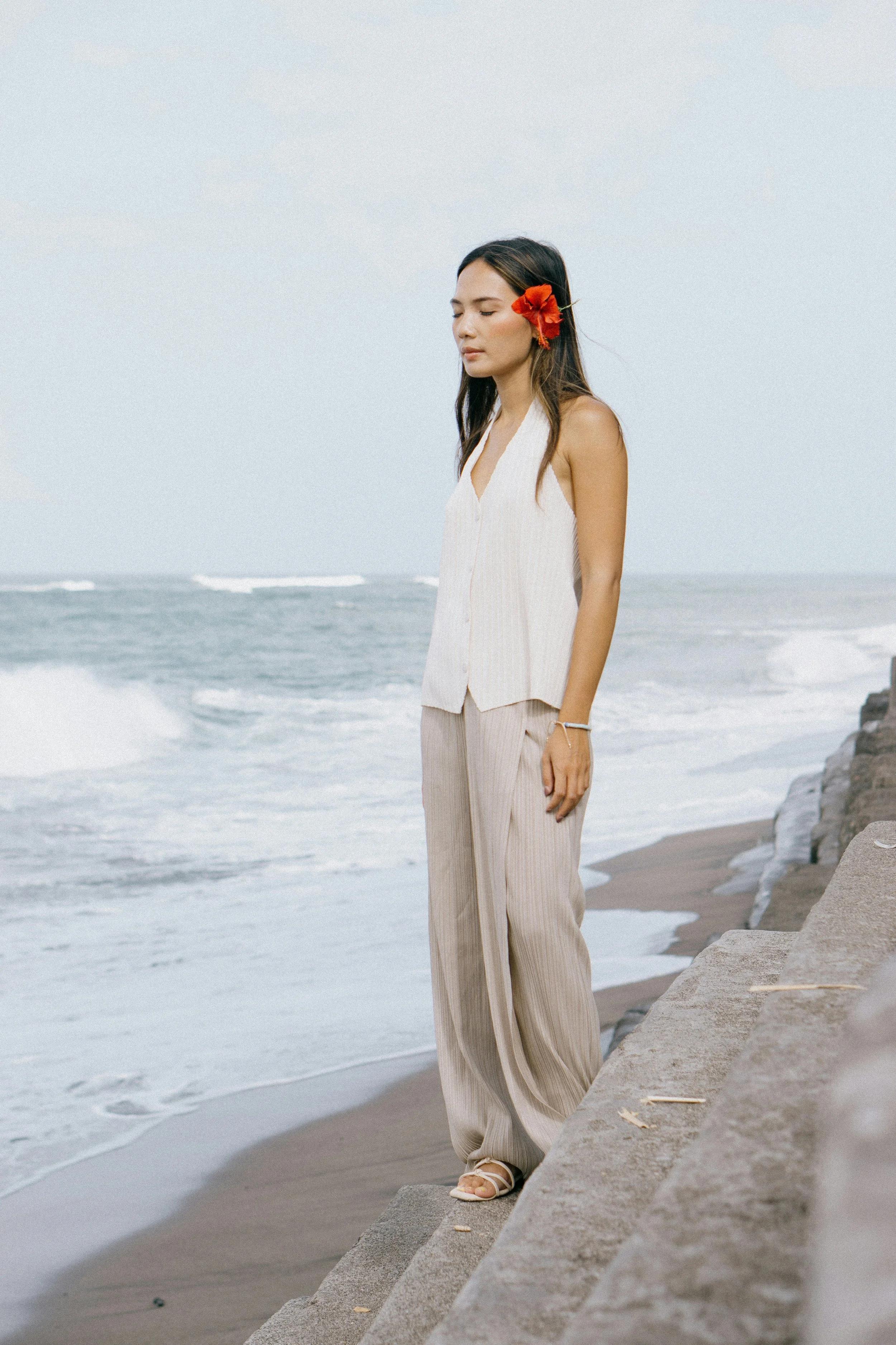 A woman standing on a stone ledge at the beach with her eyes closed, wearing a white sleeveless top and beige striped pants, with a red flower in her hair.