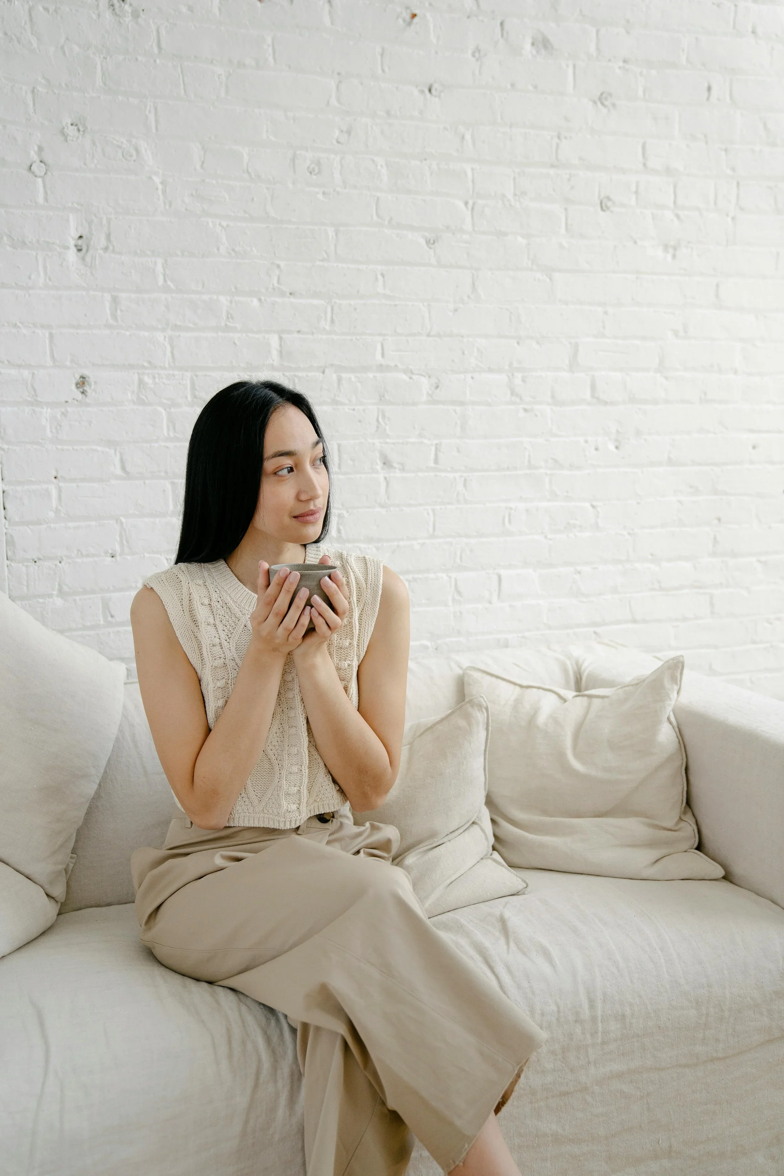 A woman sitting on a beige sofa, holding a cup with both hands, looking to her left, in front of a white brick wall.