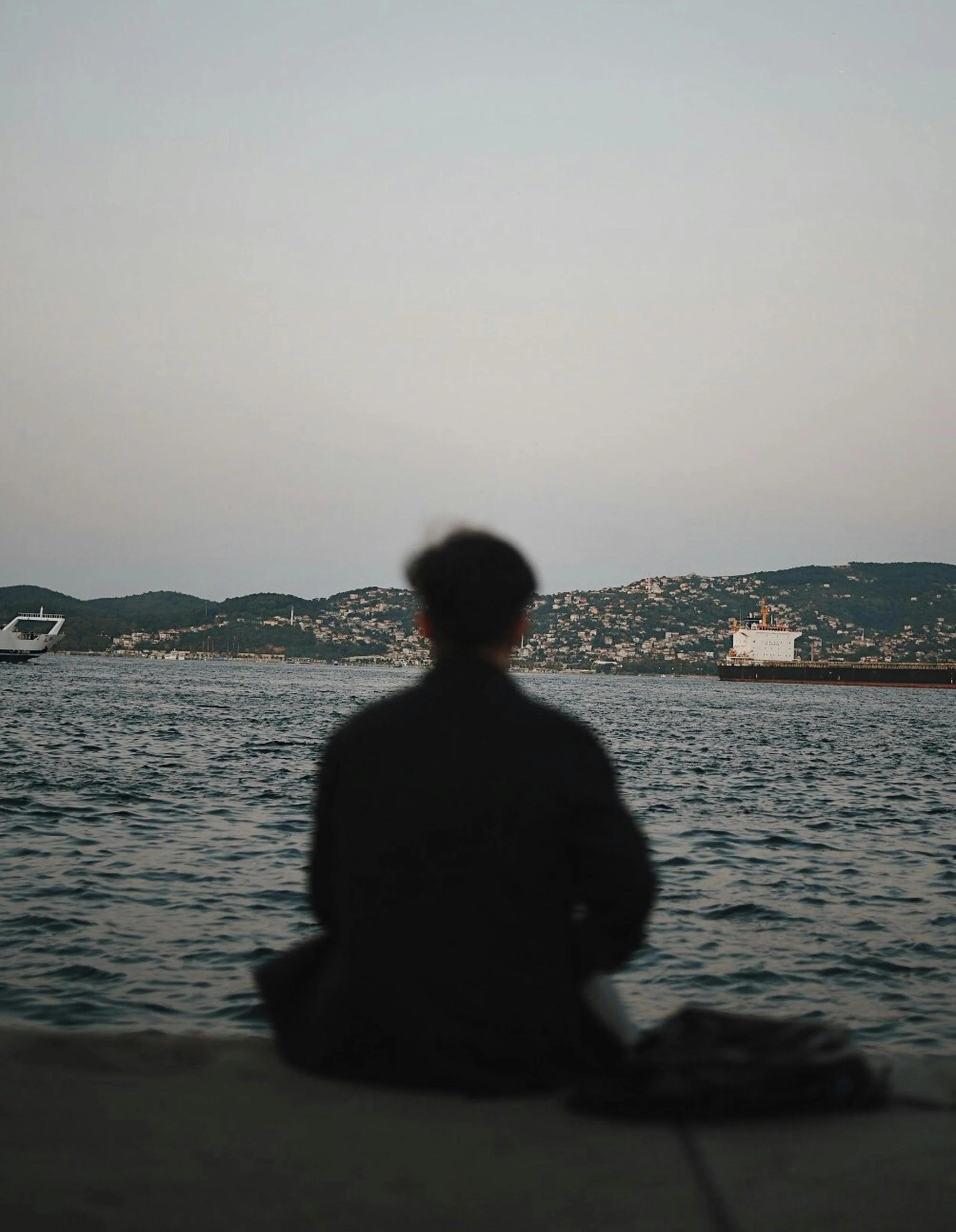 Silhouette of a person sitting on the edge of a body of water, with city buildings on hills in the background during dusk or dawn.