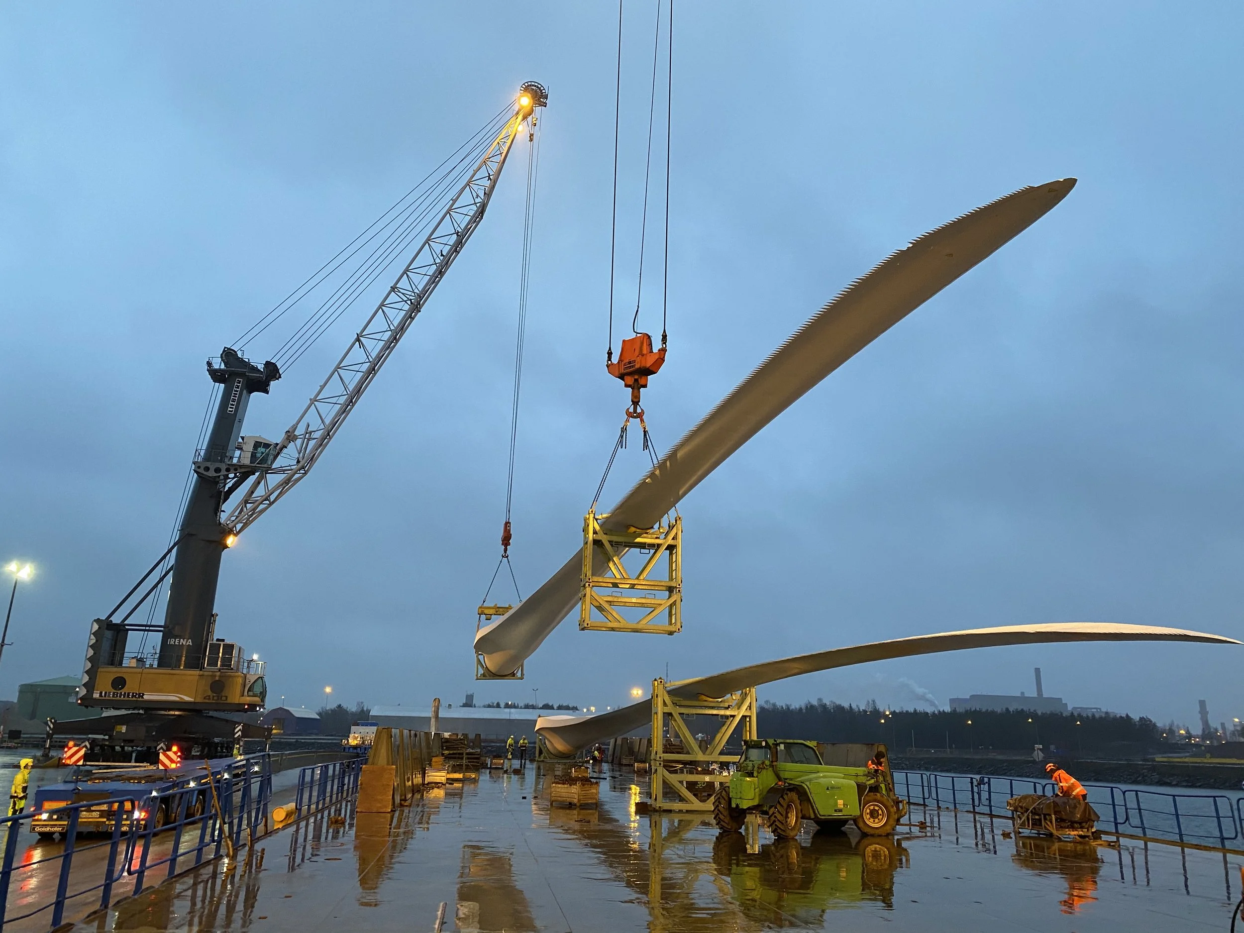Construction workers and machinery installing wind turbine blades on a platform near water, with a crane lifting a blade into place at dusk.