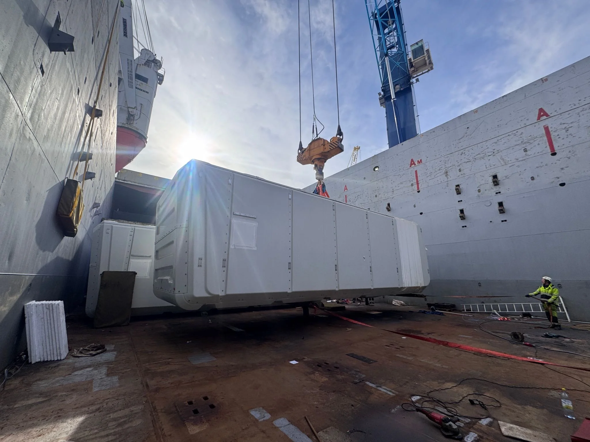 Construction workers and crane installing a large white modular unit on ship's deck at port
