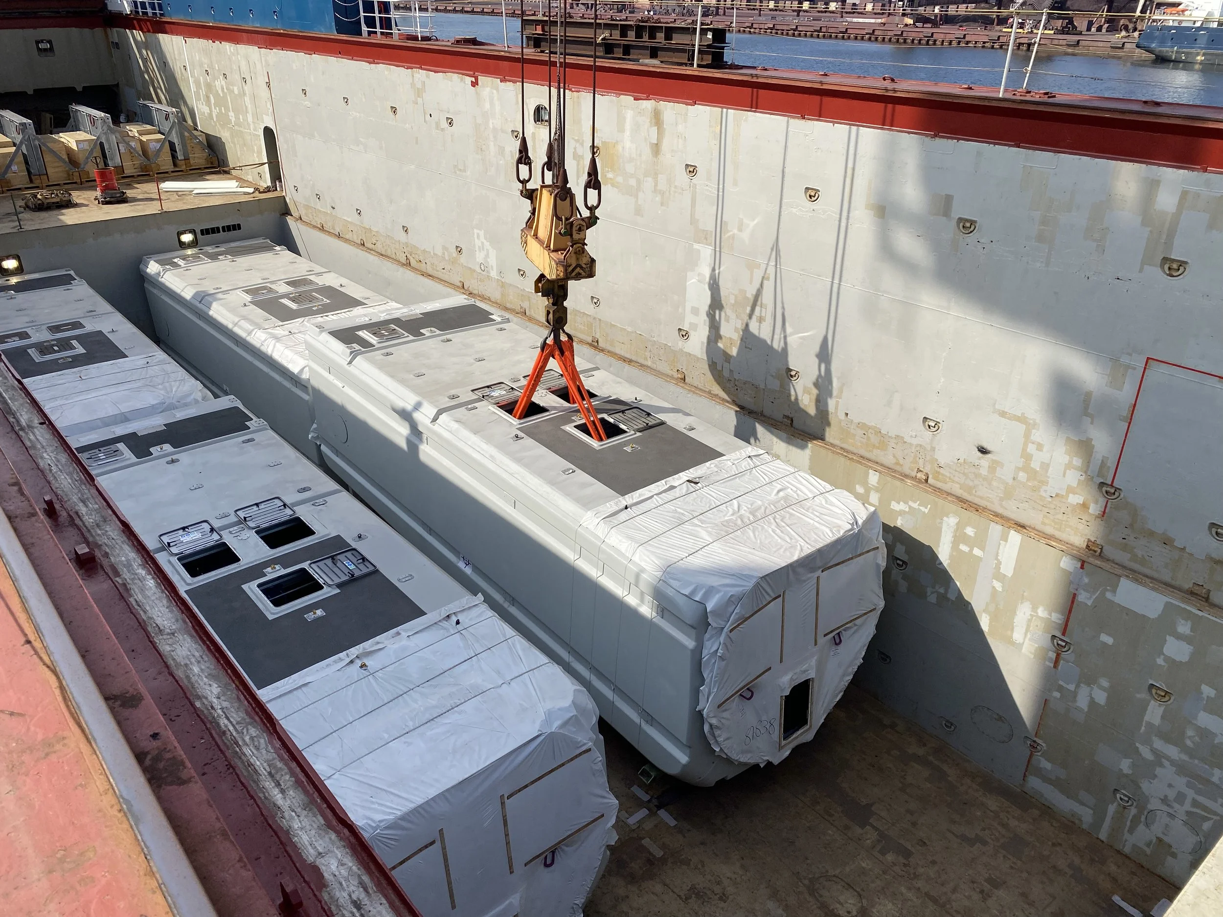 Large cargo container being lifted by a crane onto a ship's deck with several other containers wrapped in white protective covering.