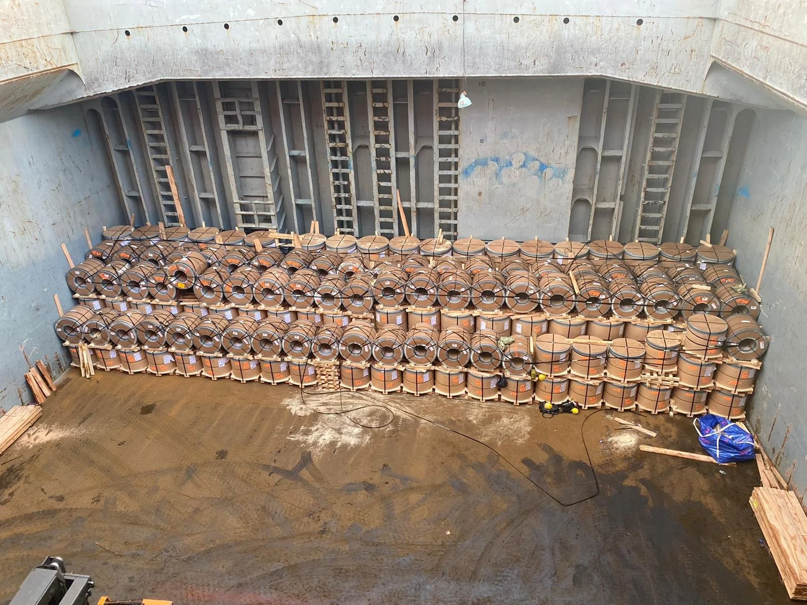 Interior view of an underground construction site with stacks of construction materials, including large rolls of wire and wooden boards, surrounded by partially finished concrete walls and ladders.
