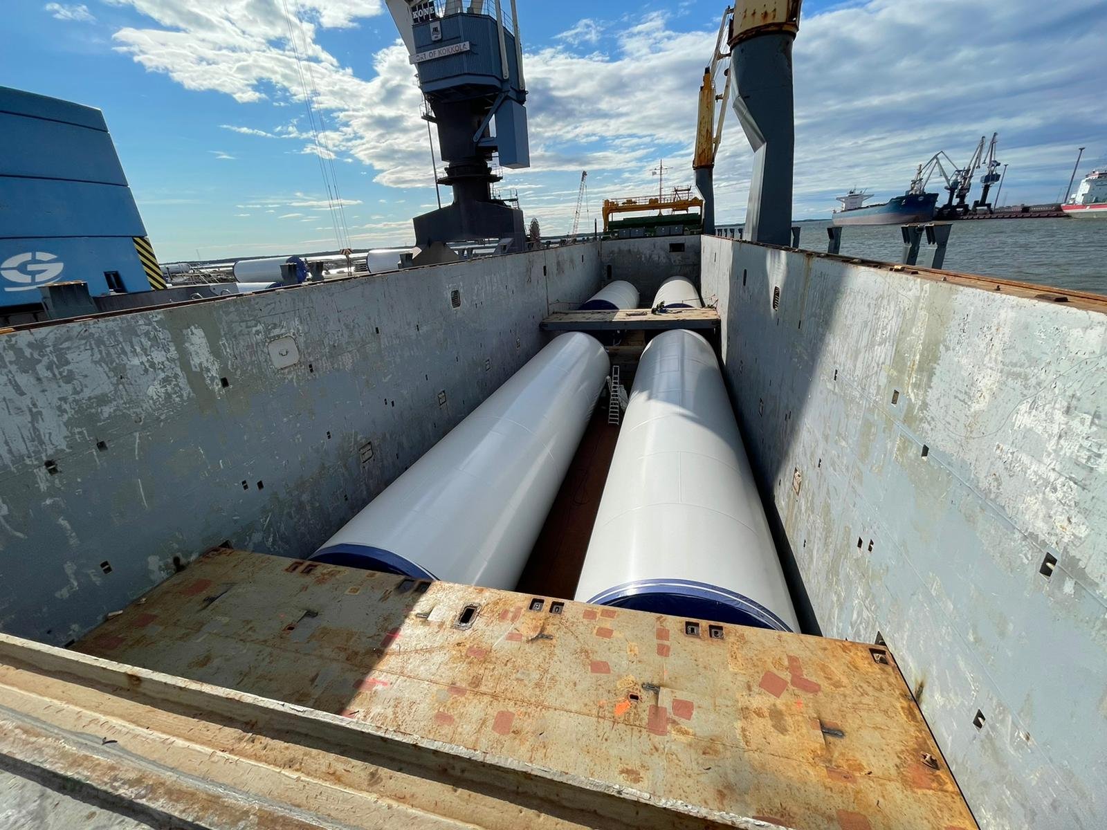 Large naval ship with two wind turbine bodies, with cranes and dockyard in background.