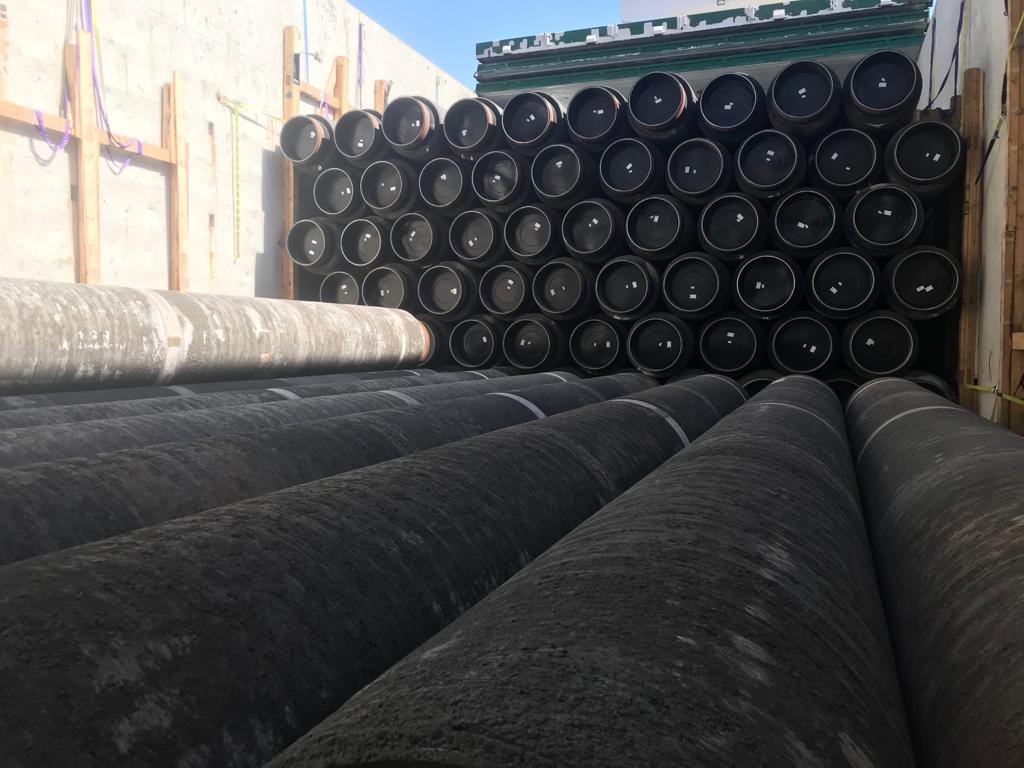Cargo ship with large black pipes and stacked black pipe caps against wooden framing under a blue sky.