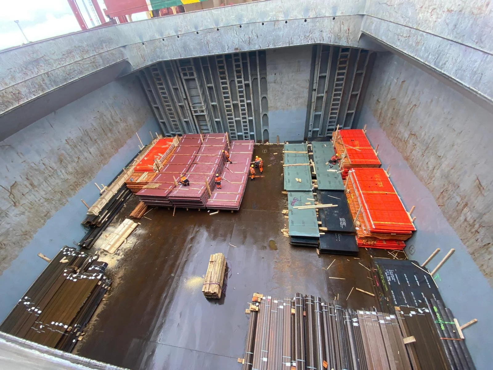 Workers assembling platforms and materials inside a large ship with concrete walls and steel reinforcement.