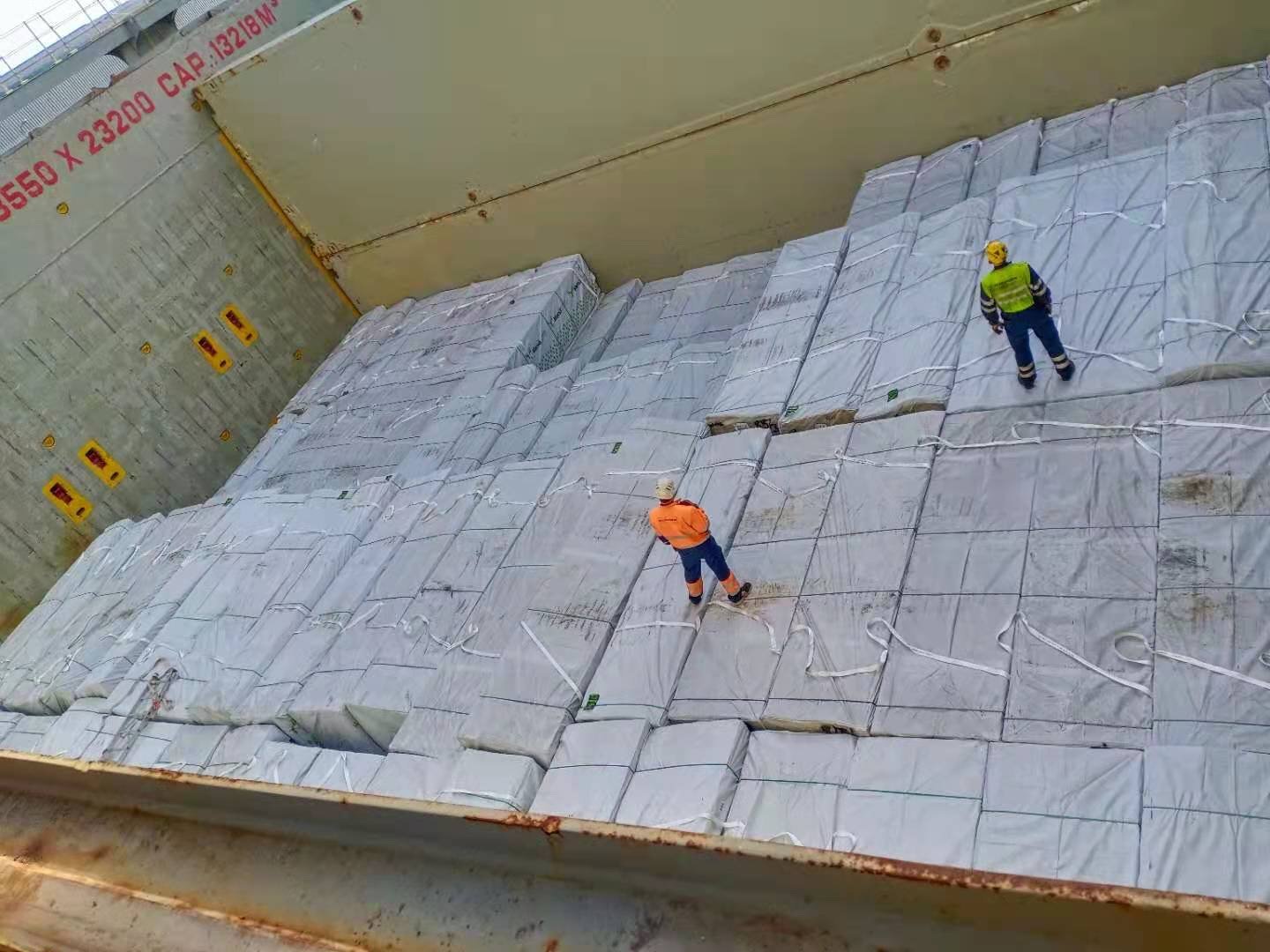Three workers on a shipping container floor, inspecting large packed cargo covered with white protective sheets, with industrial markings on the container walls.