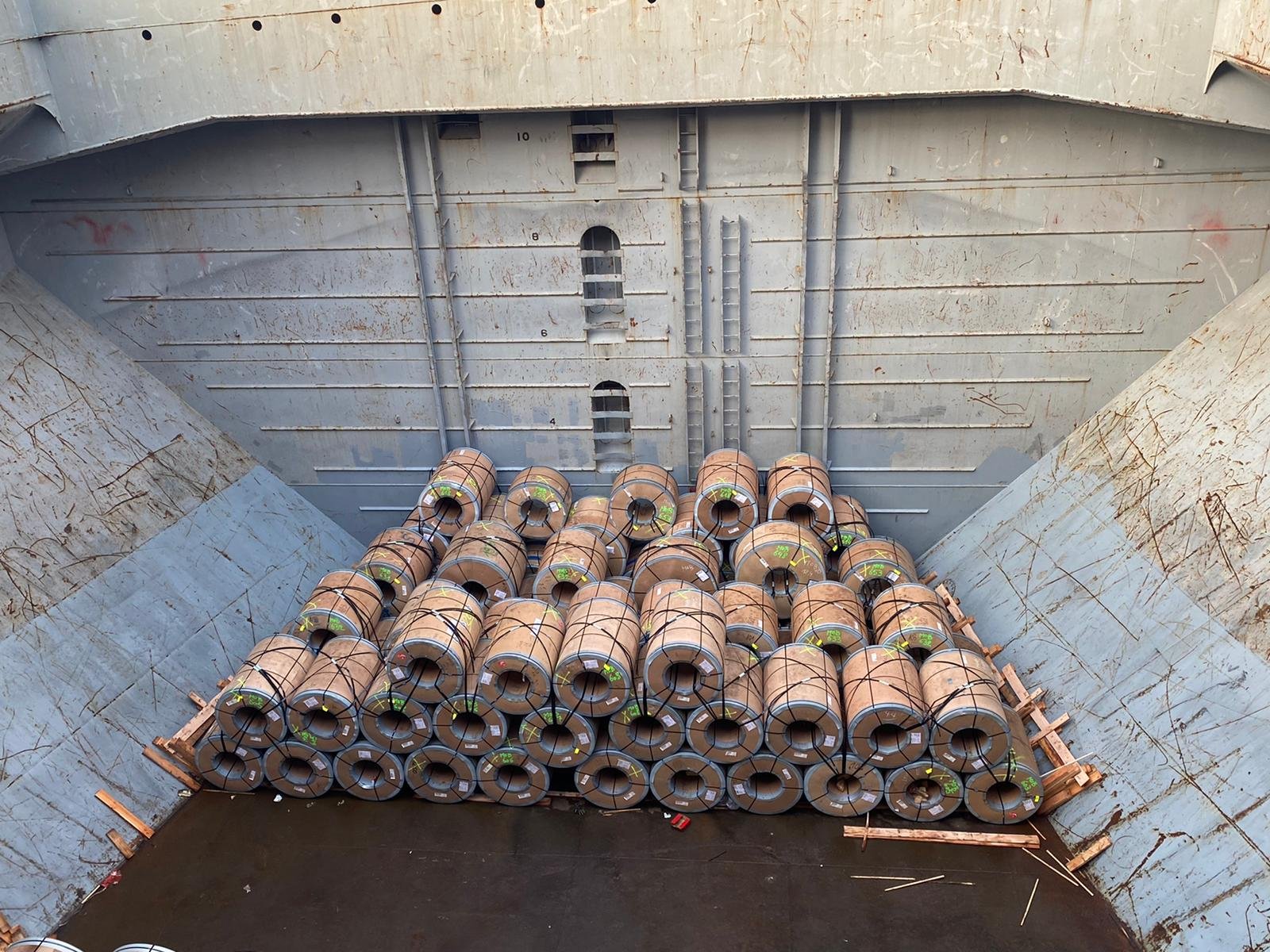 Cargo ship's hold filled with stacked large metal coils or rolls, secured with straps, viewed from above.