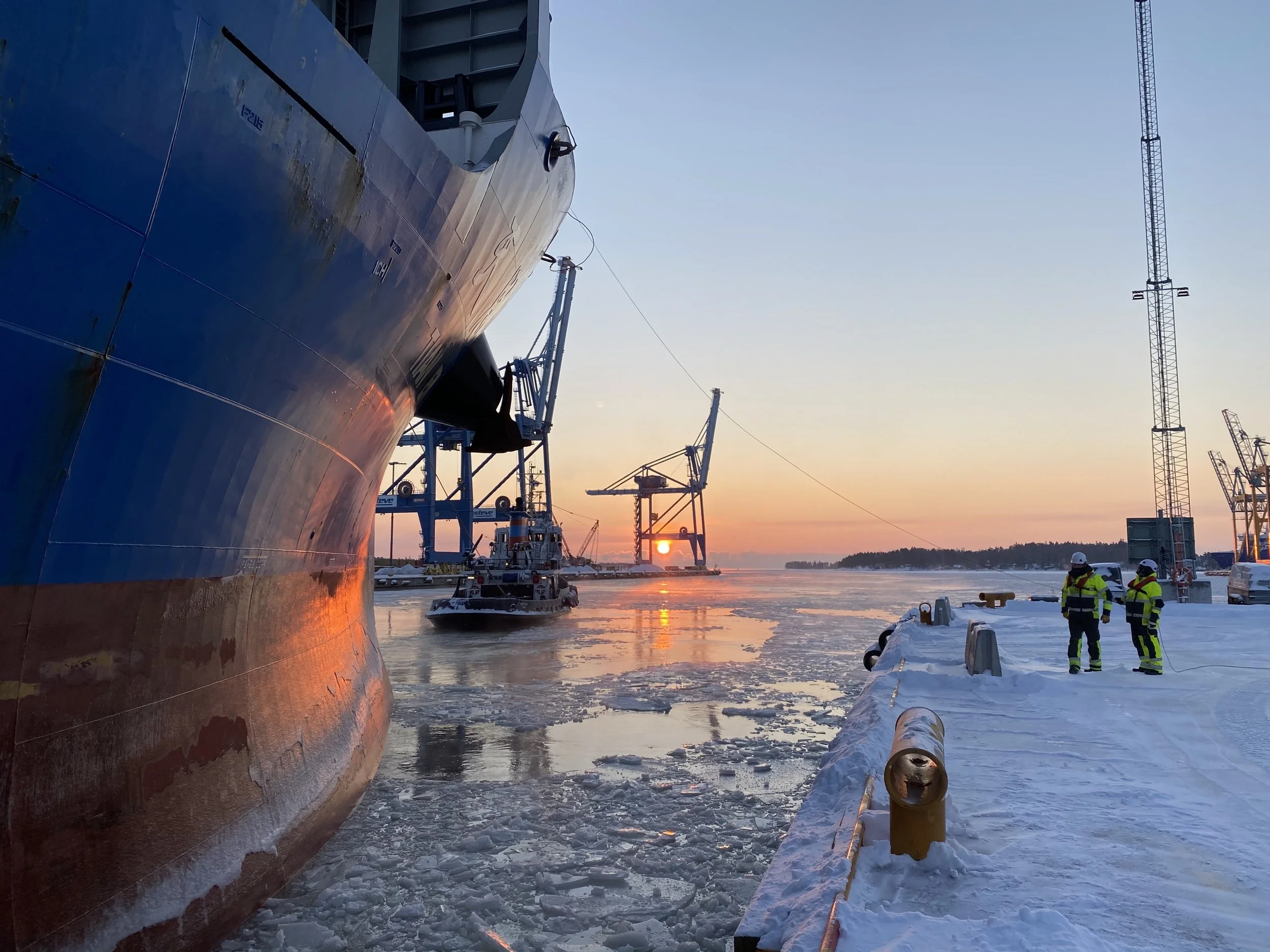 A large ship docked at a snowy harbor during sunset with two workers in safety gear standing on the snow-covered dock, and industrial cranes and a boat in the background.