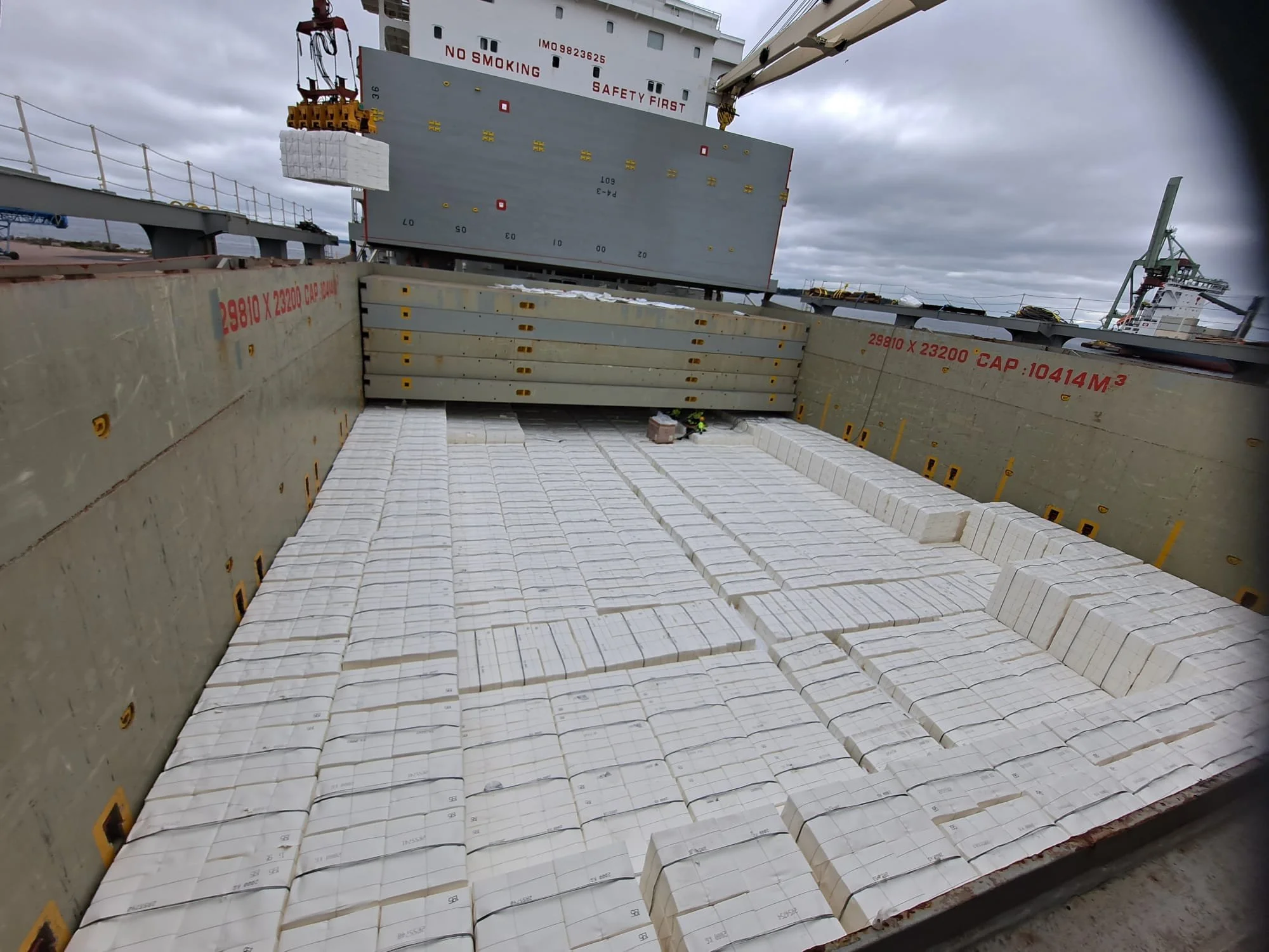 A cargo ship's hold filled with white rectangular packages, with cranes and containers visible in the background under cloudy skies.