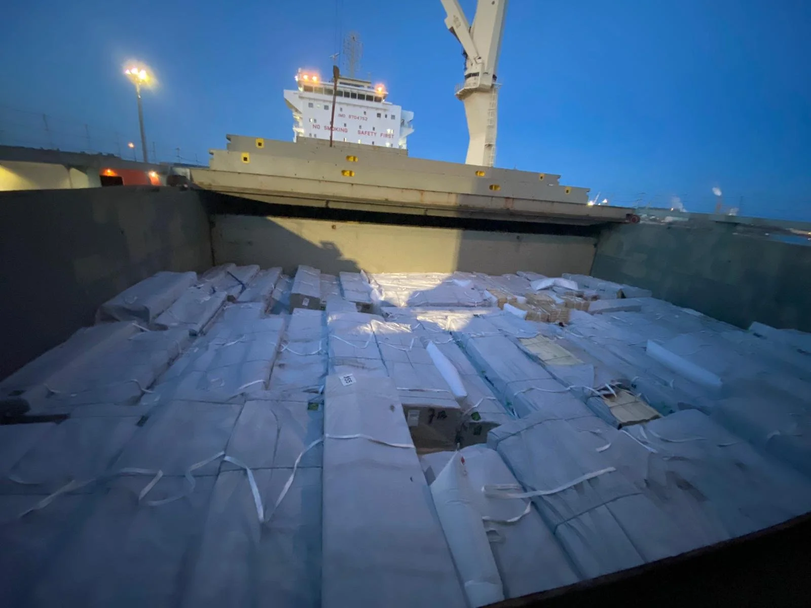 Cargo ship filled with boxes and packages, docked at port with a crane overhead during dusk.