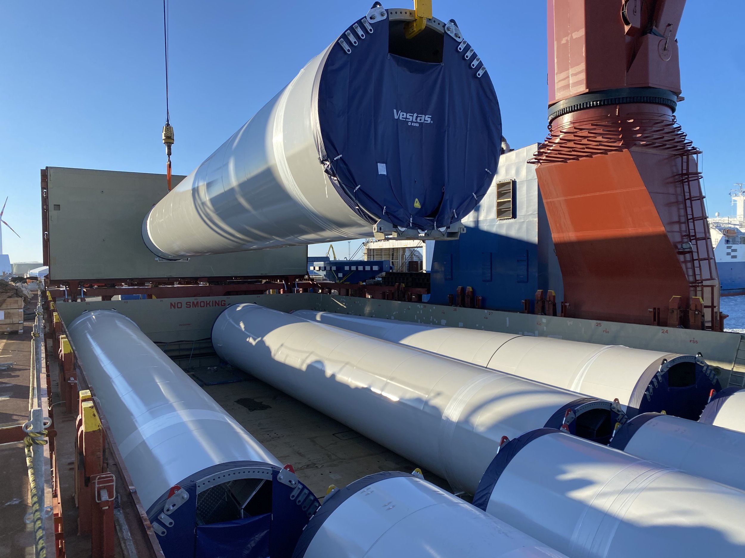 Large cylindrical wind turbine blades being loaded onto a ship, with one blade already secured and others laying on the deck, under a clear blue sky.