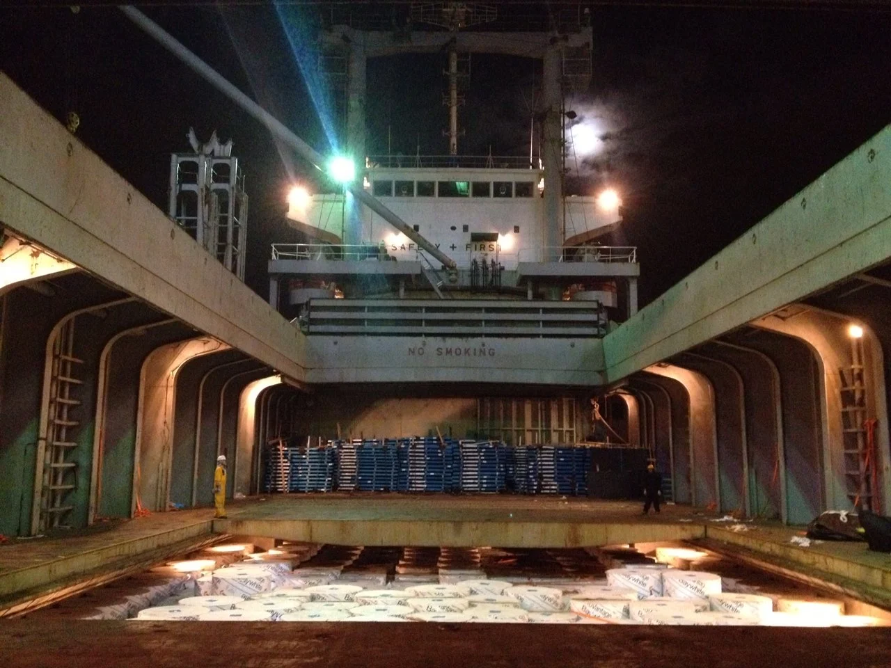 View of the deck of a large cargo ship at night, showing the ship's superstructure, crew members, and cargo on the deck, illuminated by lights under a moonlit sky.