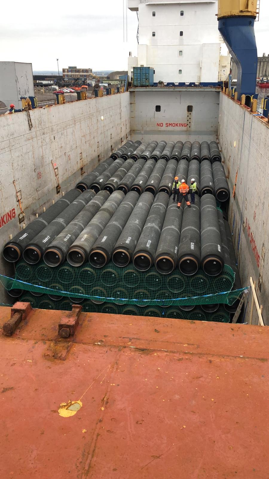 A large cargo ship's hatch filled with numerous black industrial pipes stacked horizontally. Several workers in safety gear are inspecting the pipes inside the ship.
