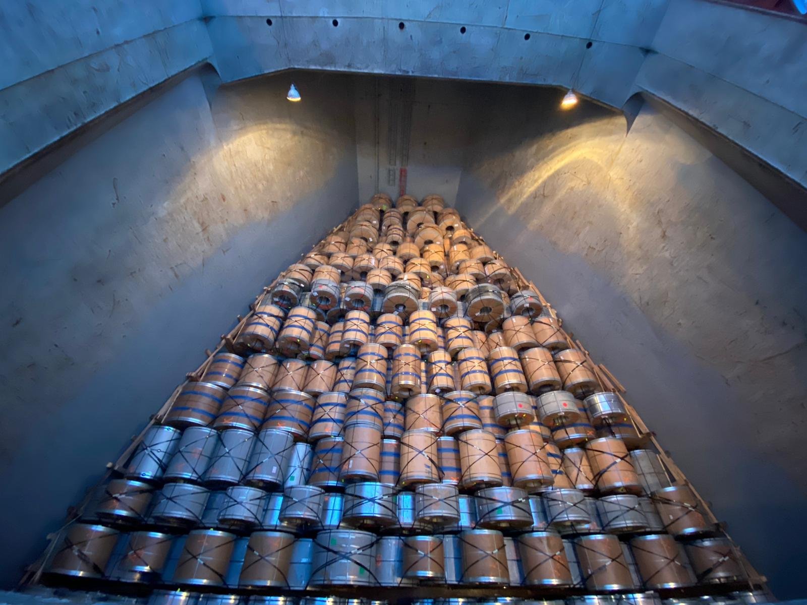 A large collection of metallic barrels stacked in a pyramid shape inside a cargo shipment.