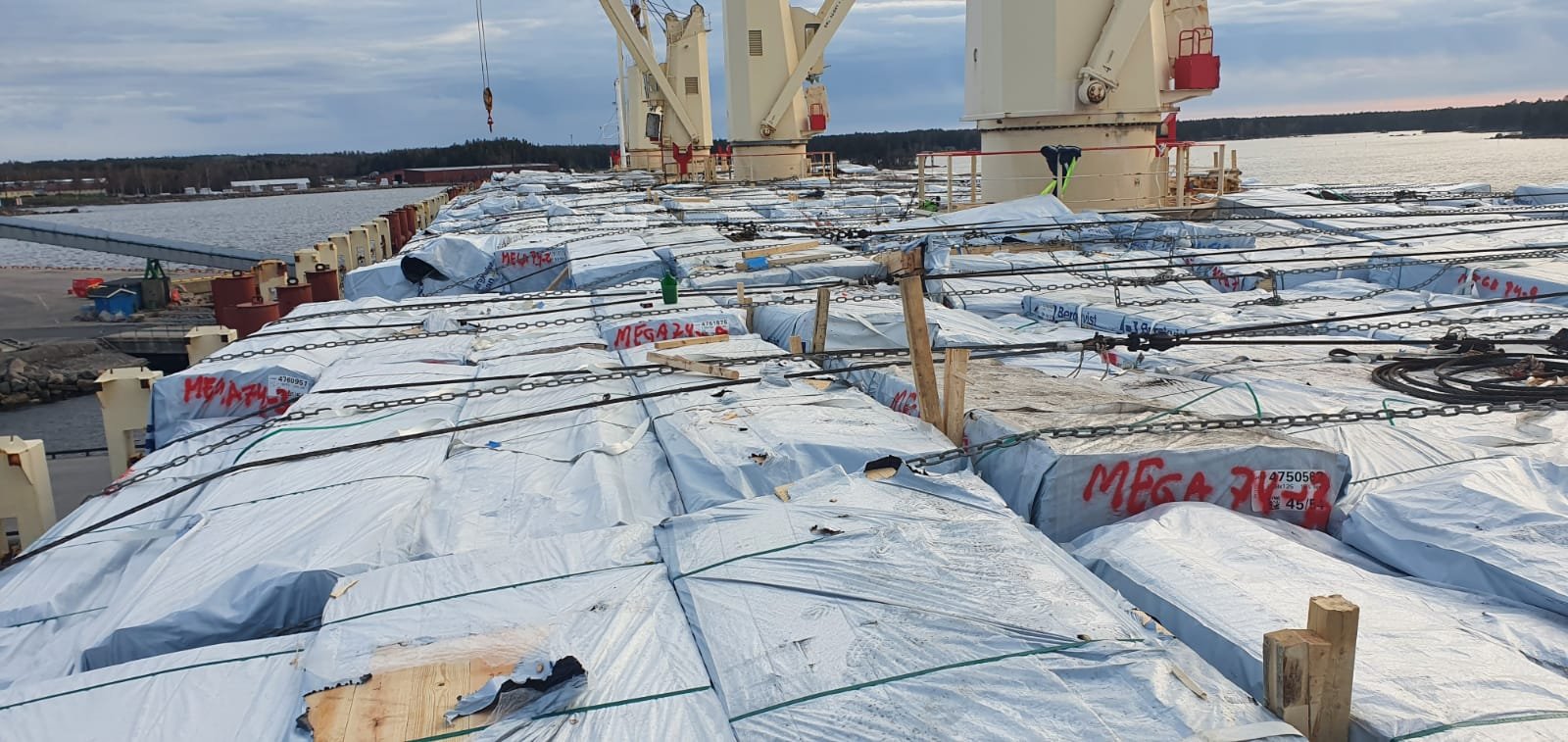 Large industrial crane on a barge on a body of water, with stacks of wrapped materials and wooden supports on the barge, and a distant shoreline with trees and buildings.