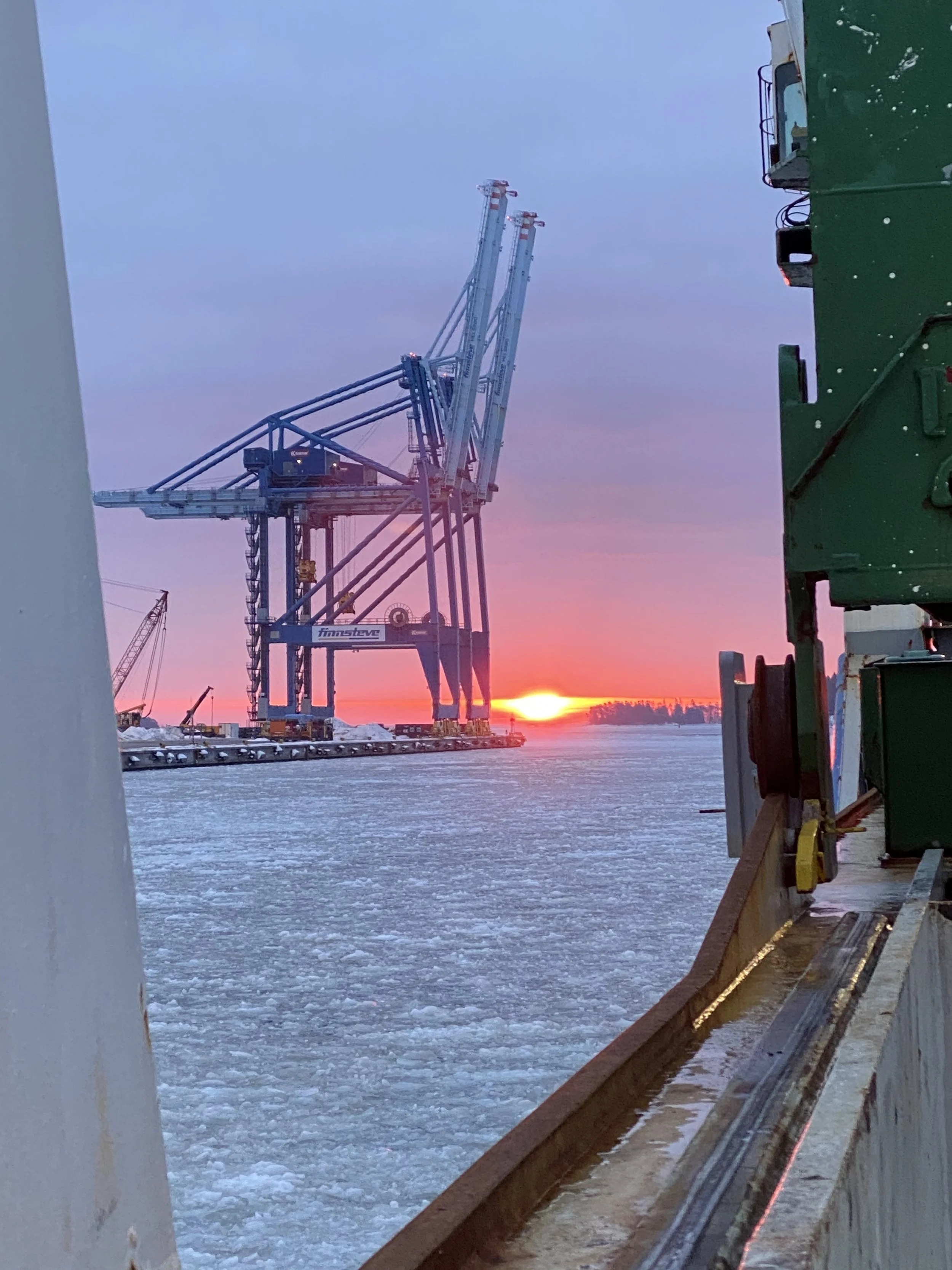 An industrial port scene at sunset with large cranes over a frozen body of water.