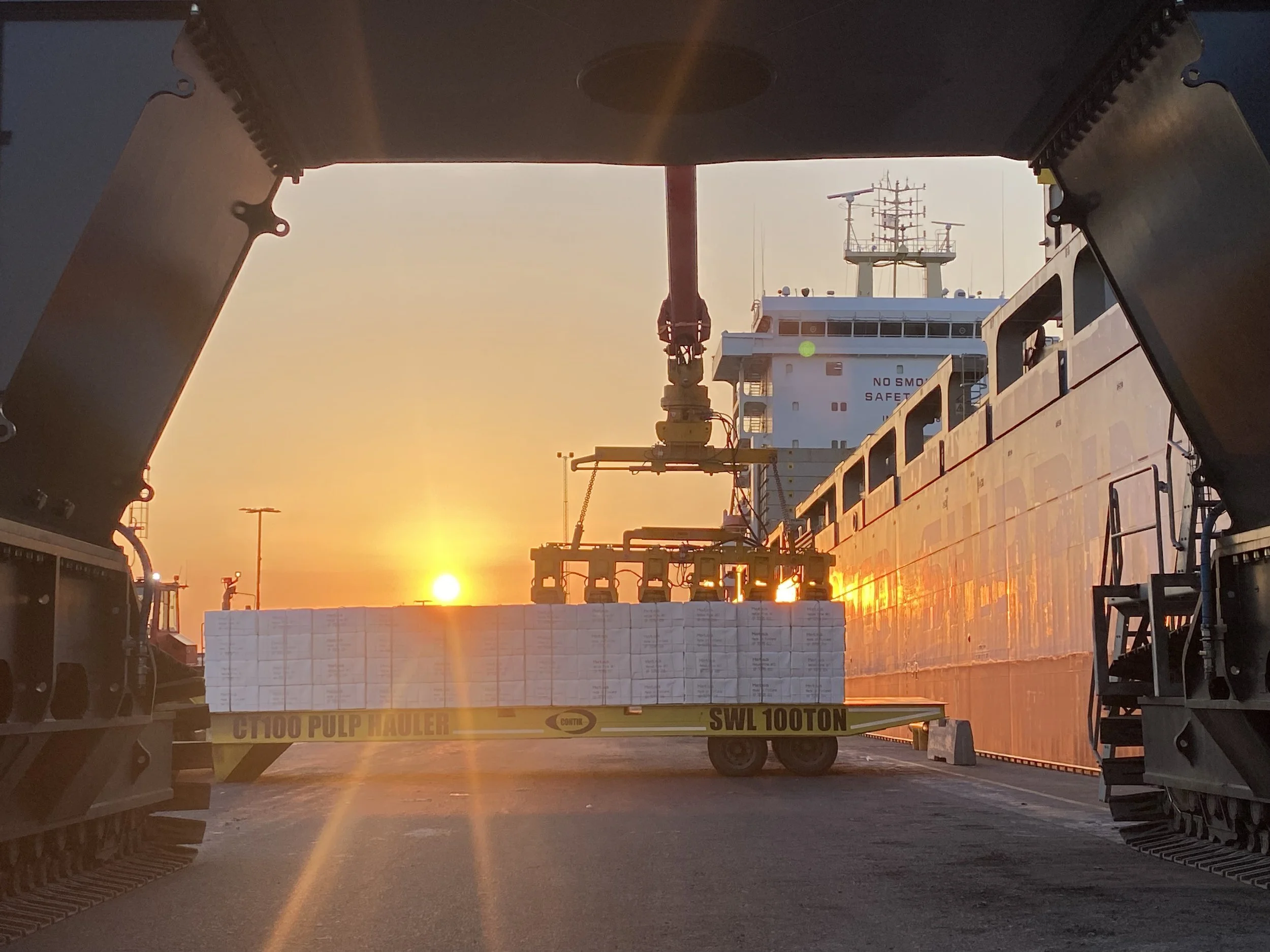 View of a cargo ship at sunset seen from through a large excavator scoop with a pile of stacked boxes being moved by a crane on the ship.