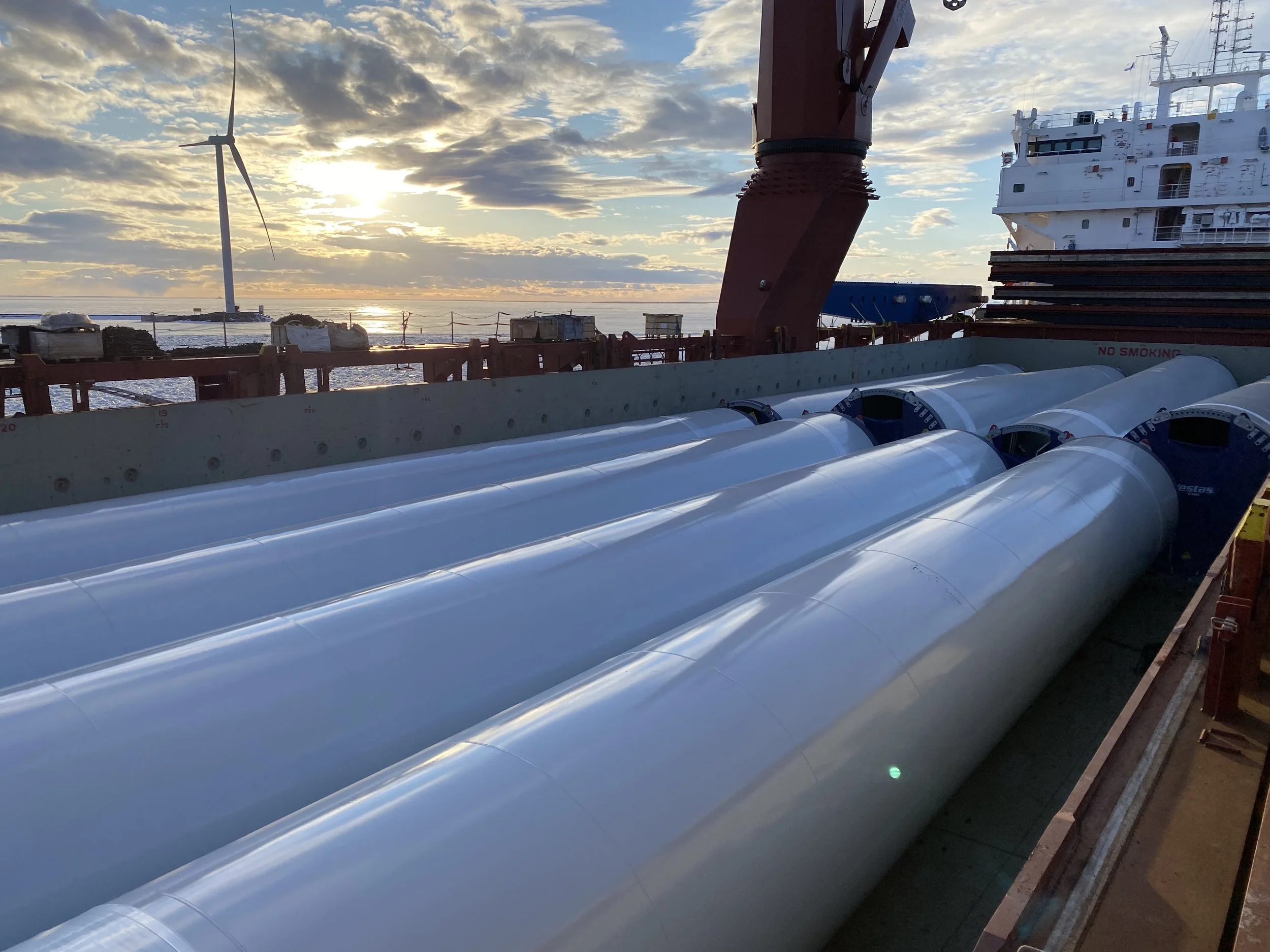 Large white wind turbine blades stored on a ship's deck with a sunset and a wind turbine in the background.