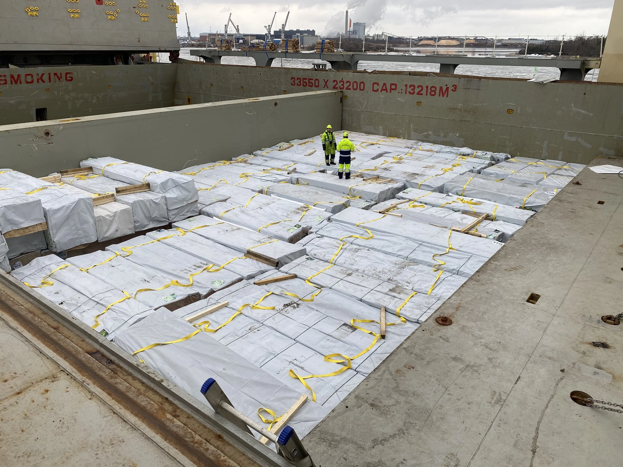 Construction workers inspecting large packages wrapped in white on a ship's deck, with industrial buildings and cranes in the background.