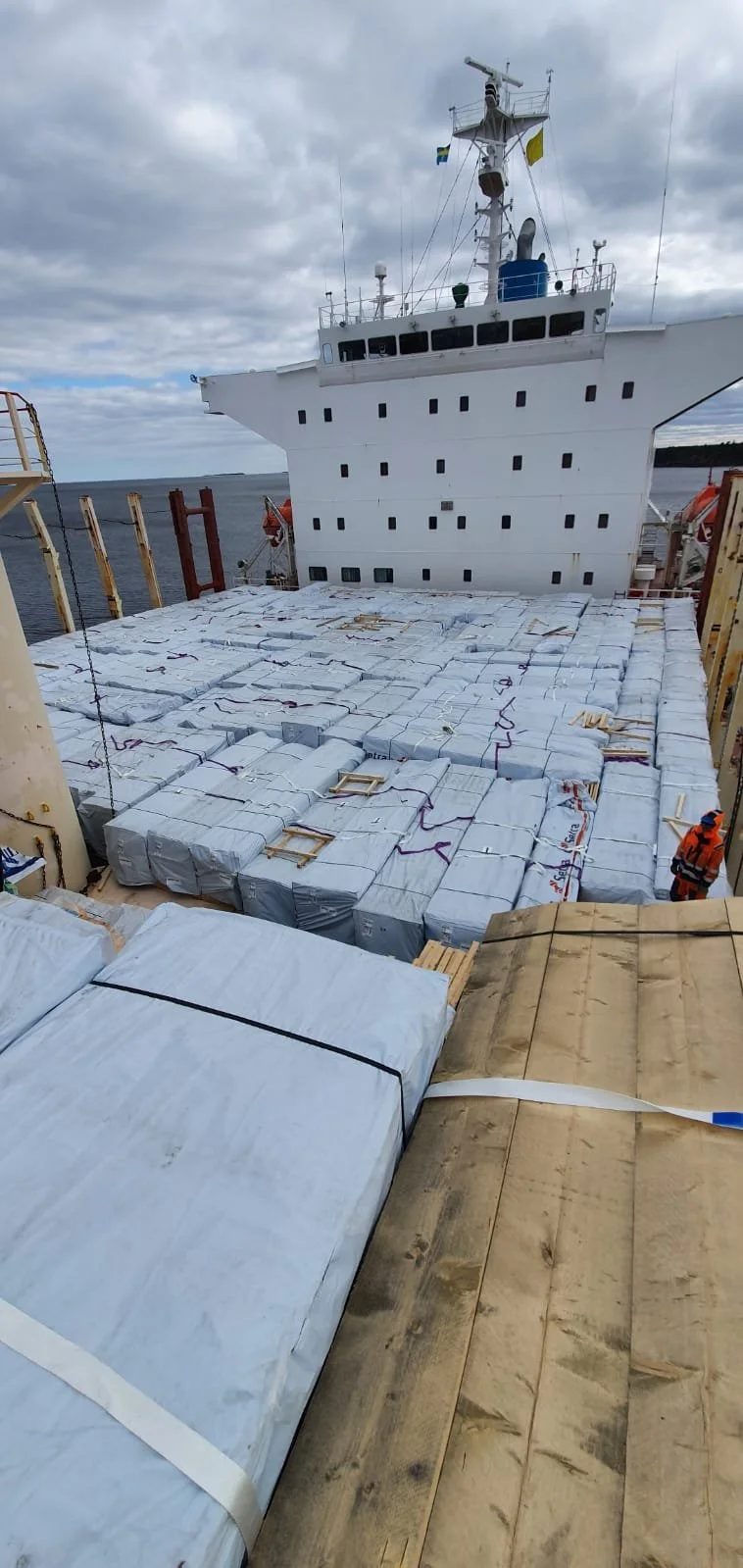 Cargo ship deck loaded with large wrapped packages, with the ship's bridge and antenna visible in the background, over a cloudy sky and water.