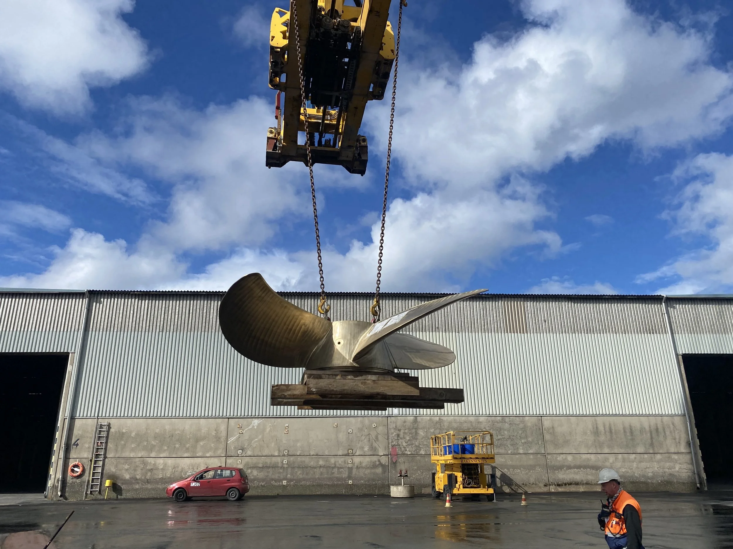Large ship propeller being lifted by a crane in an industrial building yard with a worker in safety gear, a red car, and a yellow lift in the background.