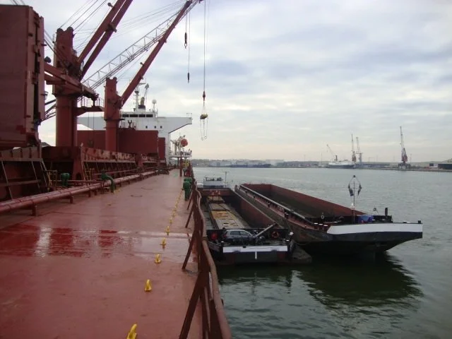 A red cargo ship docked at a port, with a large loading crane, and a barge beside it with trucks on deck, on a cloudy day.
