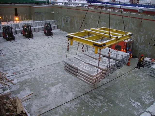 Construction site with a large yellow lifting frame hoisting metal slabs into a deep cargo ship carefully.