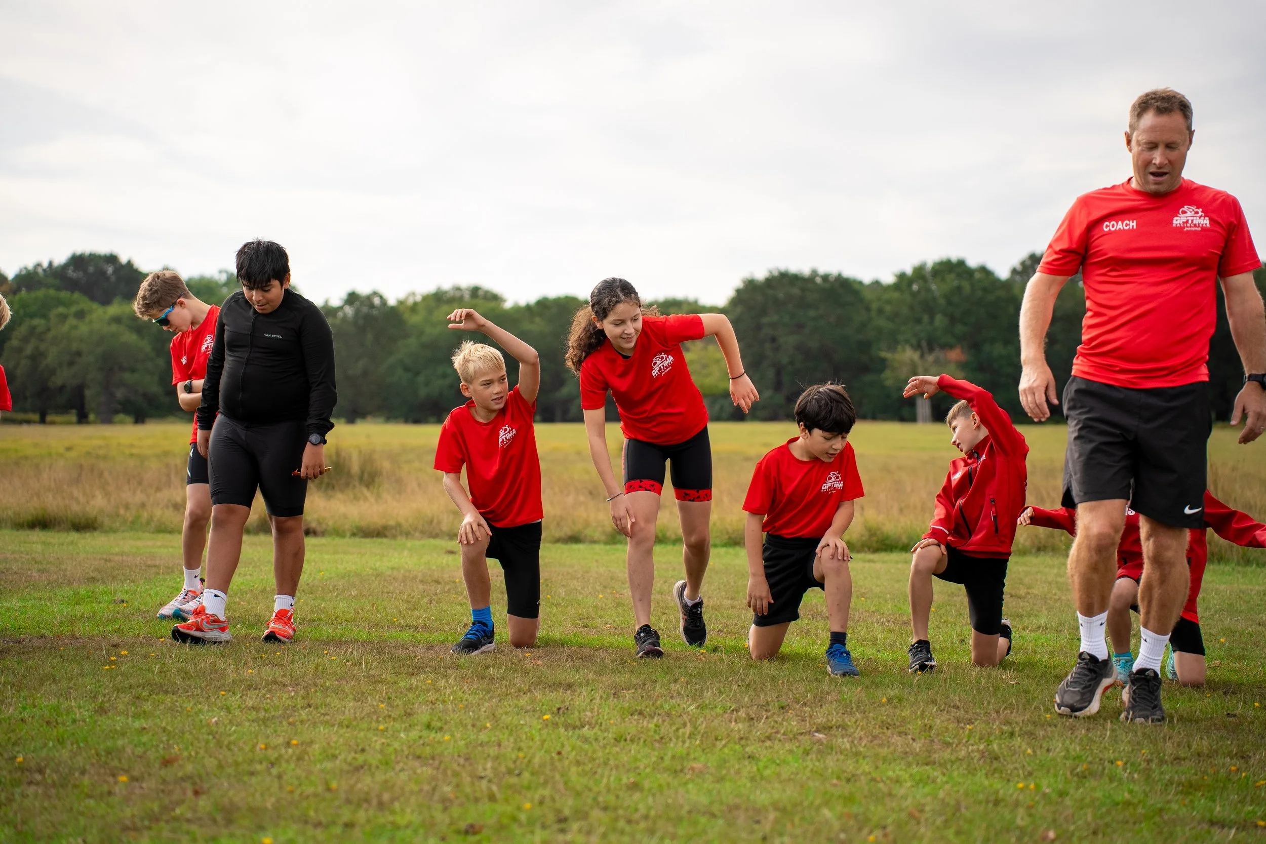 Group of children with coach Mark Klein in red shirts participating in an outdoor athletic activity on a grassy field.