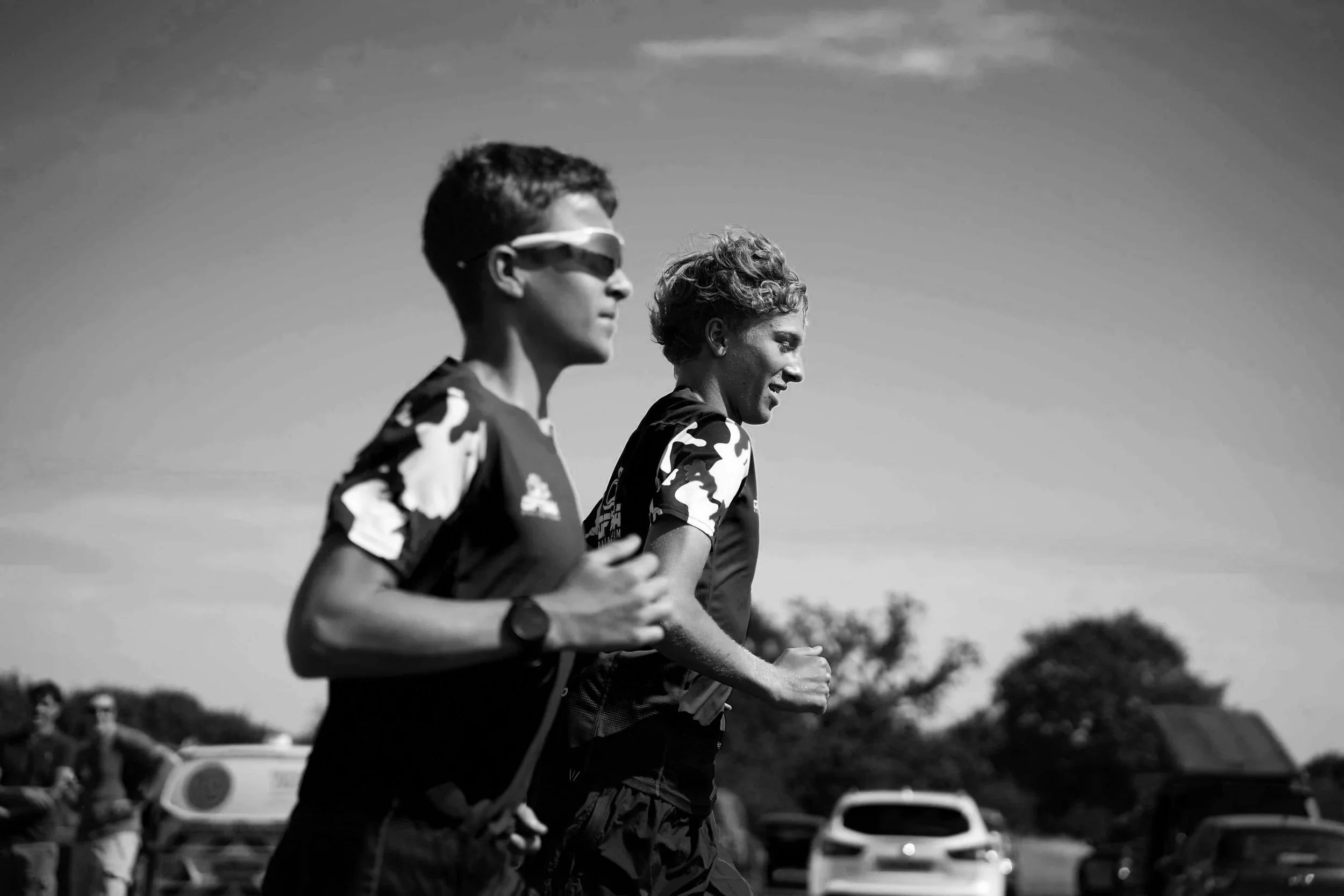 Two runners, a young boy and a woman, jogging outdoors in a race or training session, with vehicles and trees in the background.