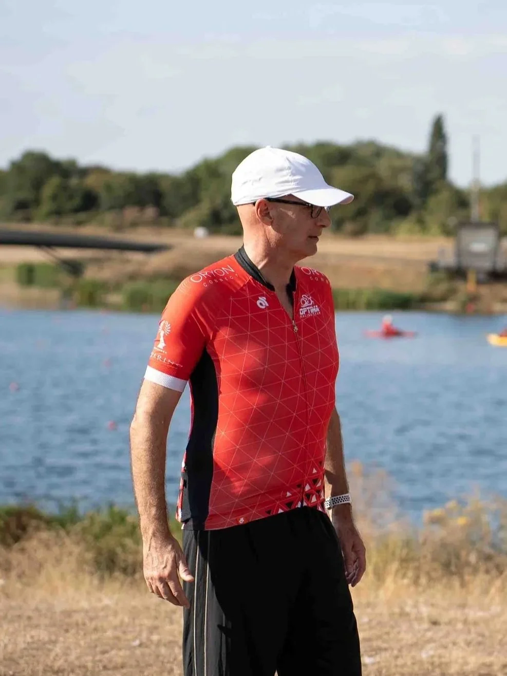 A man wearing a white cap, glasses, a red sports shirt, and black pants standing outdoors near a body of water with boats and kayaks in the background.