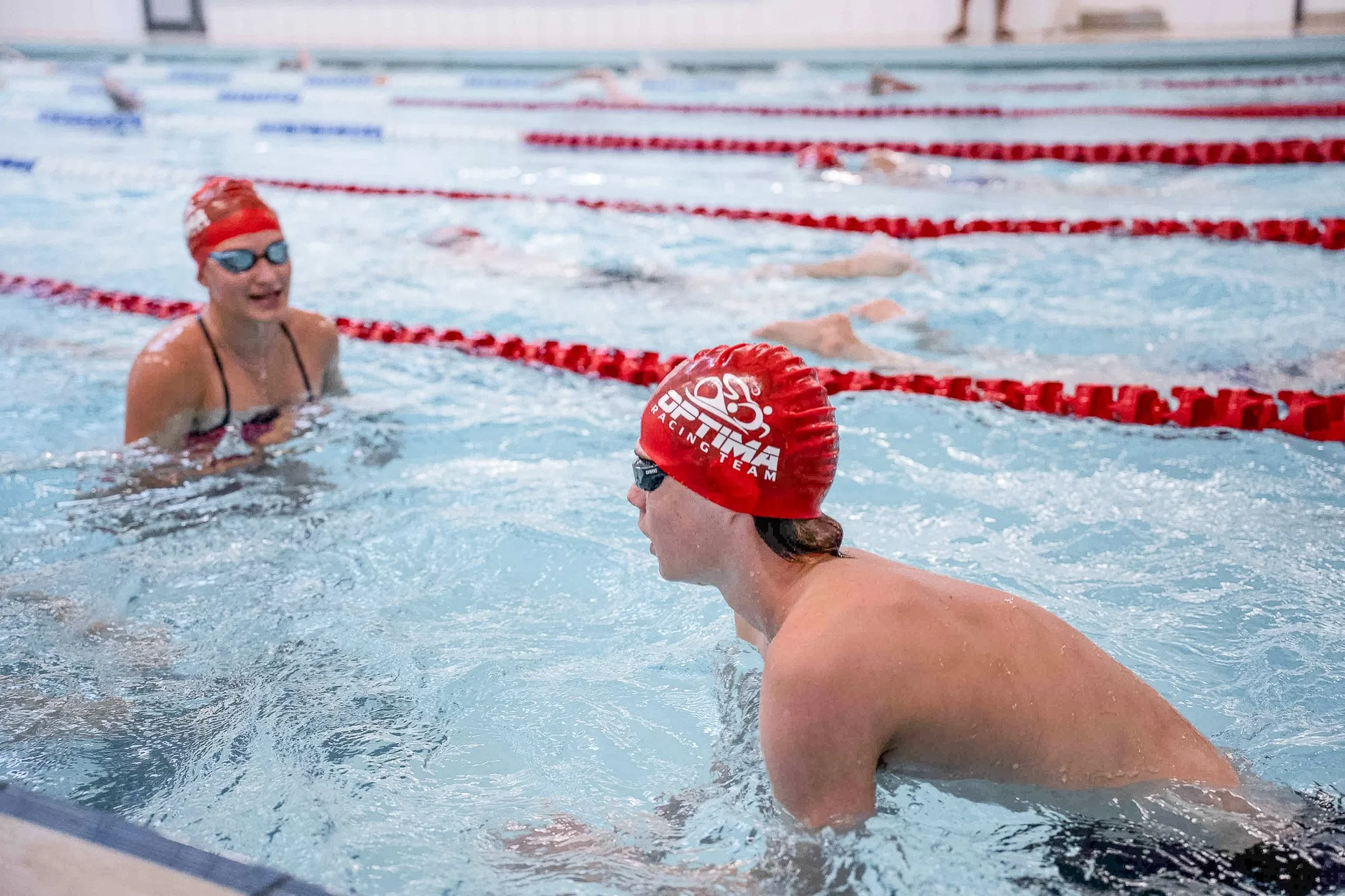 Swimmers in a pool, wearing red swim caps and goggles, with one swimmer talking to another during a swimming practice or competition.