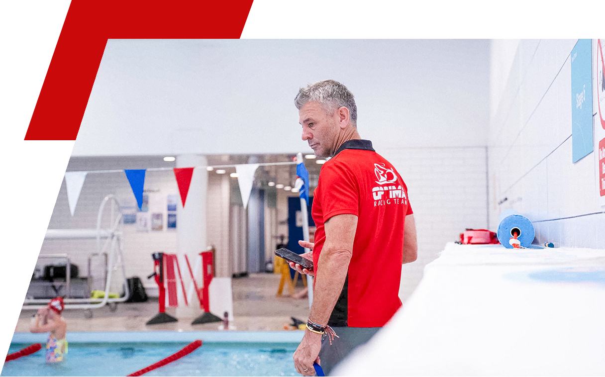 A swimming coach James Beckinsale at an indoor pool looking at his phone while a swimmer practices in the background.