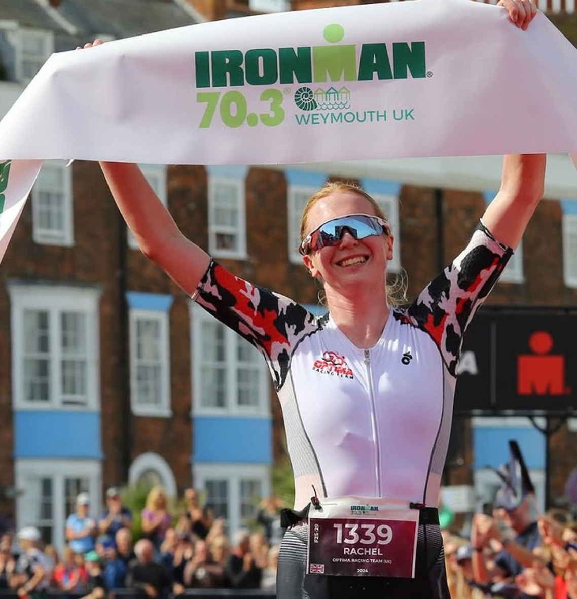 A smiling female athlete wearing sunglasses and a white cycling suit with red, black, and pink camouflage sleeves, holding a large banner above her head at the Ironman 70.3 Weymouth UK race, with a crowd of spectators and brick buildings in the background.