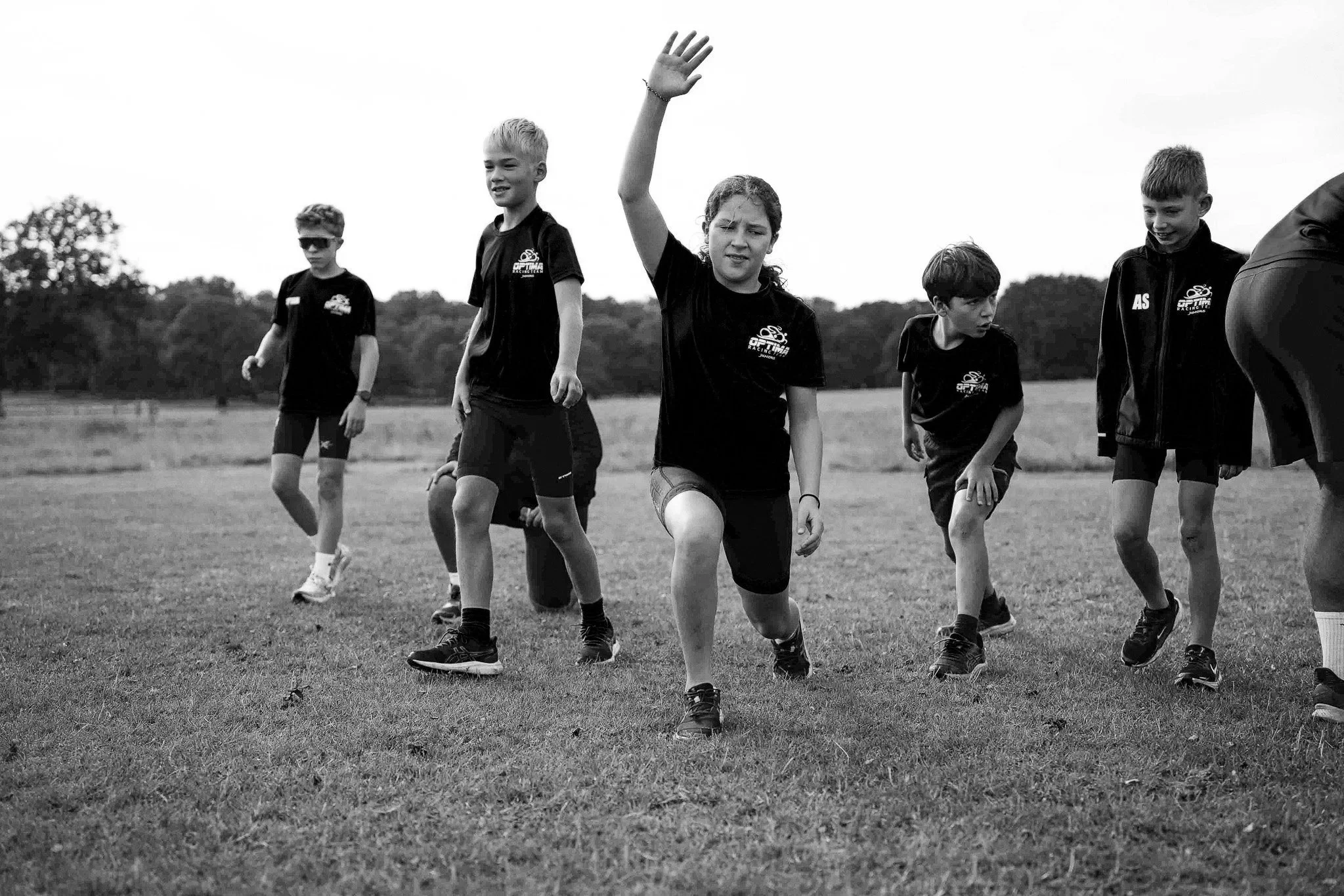 Children wearing athletic clothing are running and playing on a grassy field outdoors, with trees in the background.
