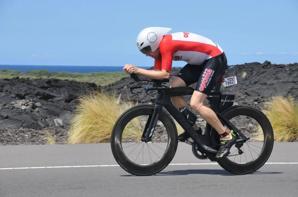A person wearing a red and white cycling suit and a white helmet rides a black time-trial bicycle on a paved road with a rocky landscape and ocean in the background.