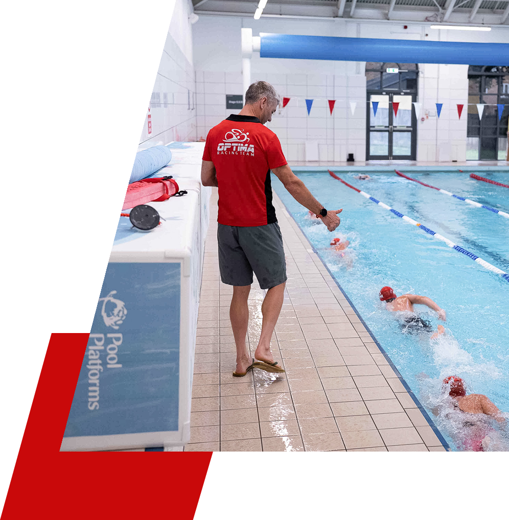 A swimming coach in a red shirt and grey shorts standing by an indoor swimming pool, giving instructions to young swimmers in the water during a swim practice or competition.