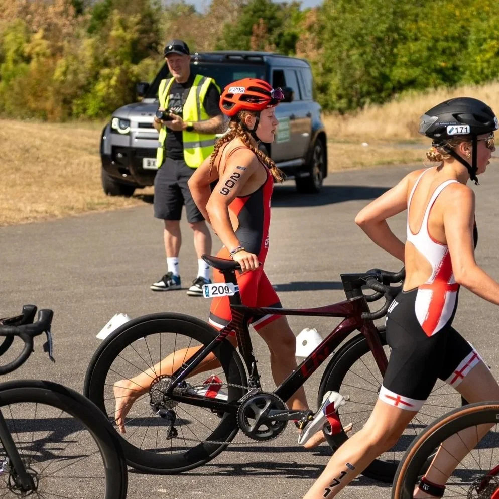 Two female triathletes with helmets and race numbers, one walking with her bike, in a transition area, with a man in a yellow safety vest and a black vehicle in the background.