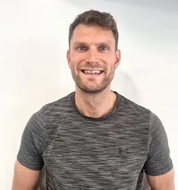 Smiling man with short brown hair and a beard, wearing a gray t-shirt, standing against a plain white background.
