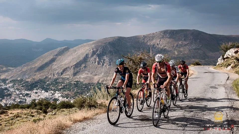 Five cyclists riding on a mountain road with hills and a cloudy sky in the background.
