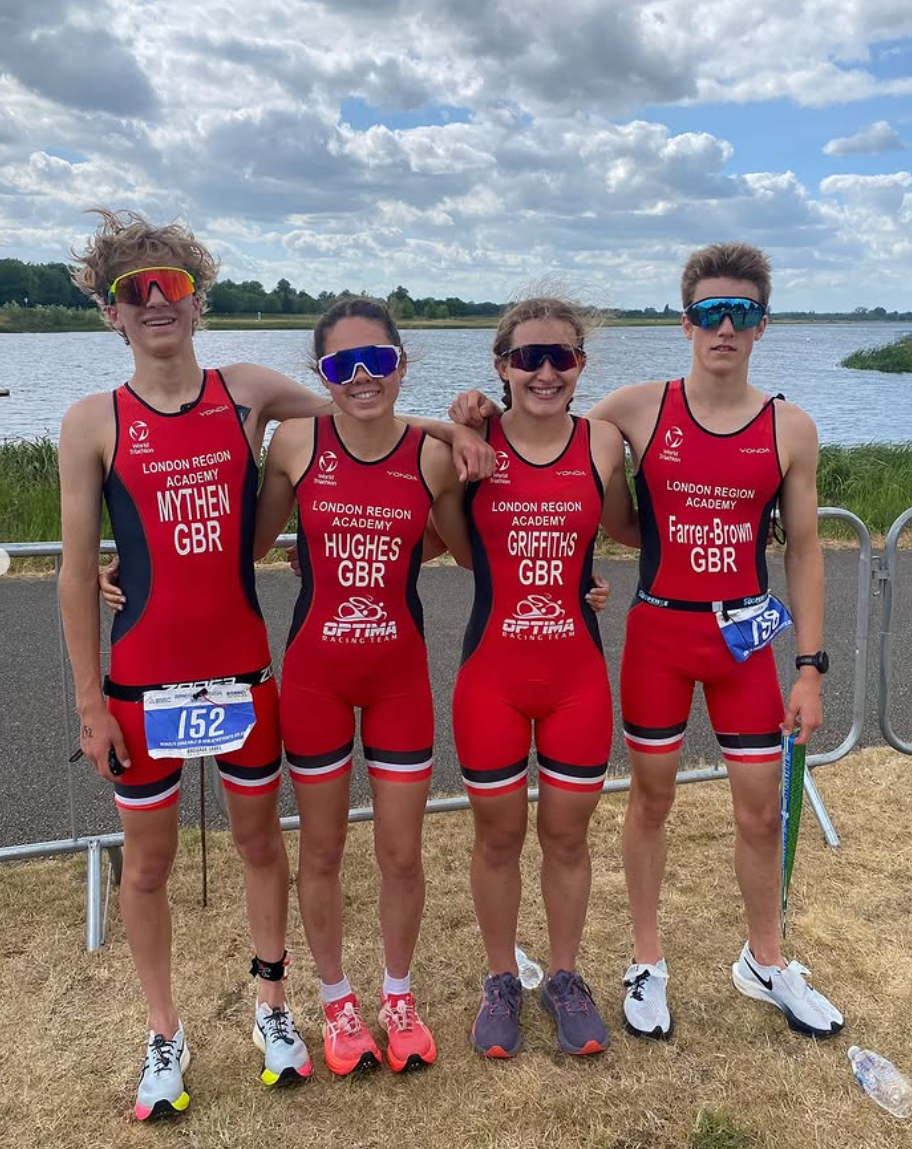 Four young athletes in red and black triathlon suits standing side by side outdoors near a lake, smiling at the camera, wearing sunglasses and athletic shoes. They are part of the London Region Academy and are from Great Britain, with one holding a water bottle, and one wearing a race bib number 152. The background features a cloudy sky and trees.