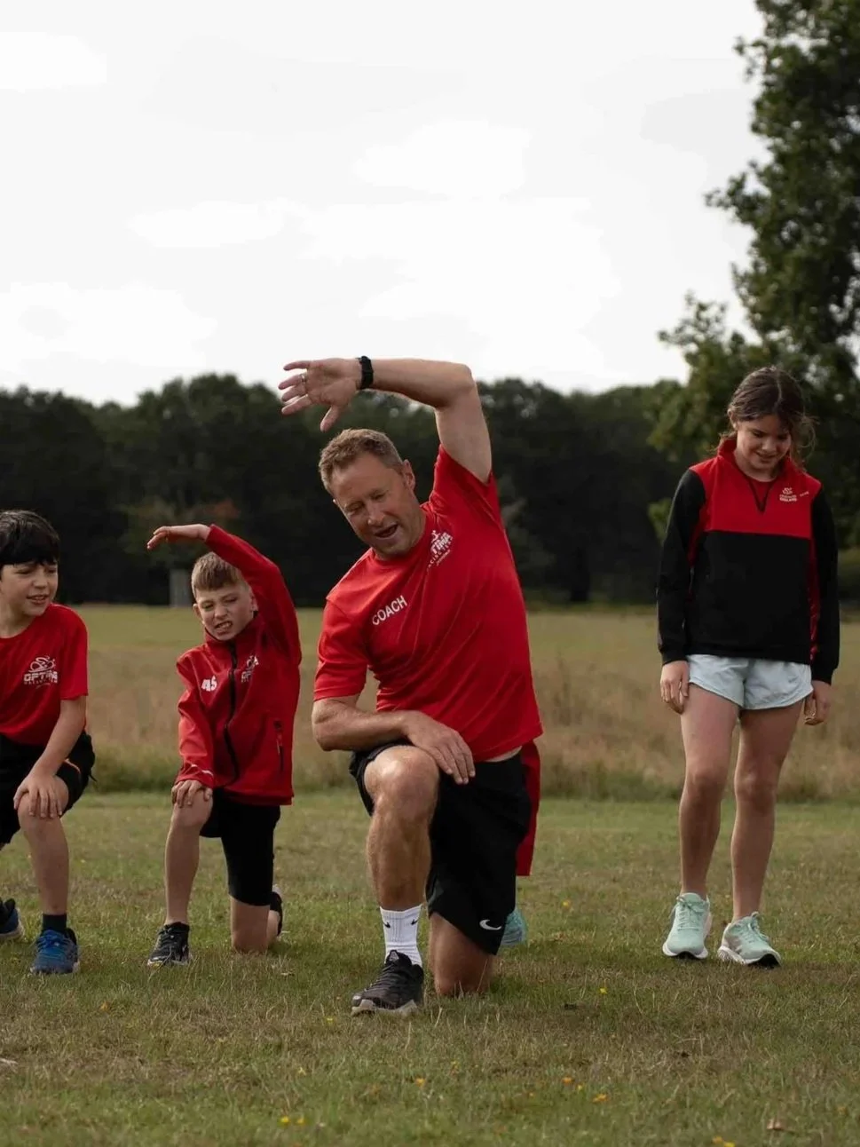 A coach and three children participating in an outdoor stretching or warm-up exercise on a grassy field with trees in the background.