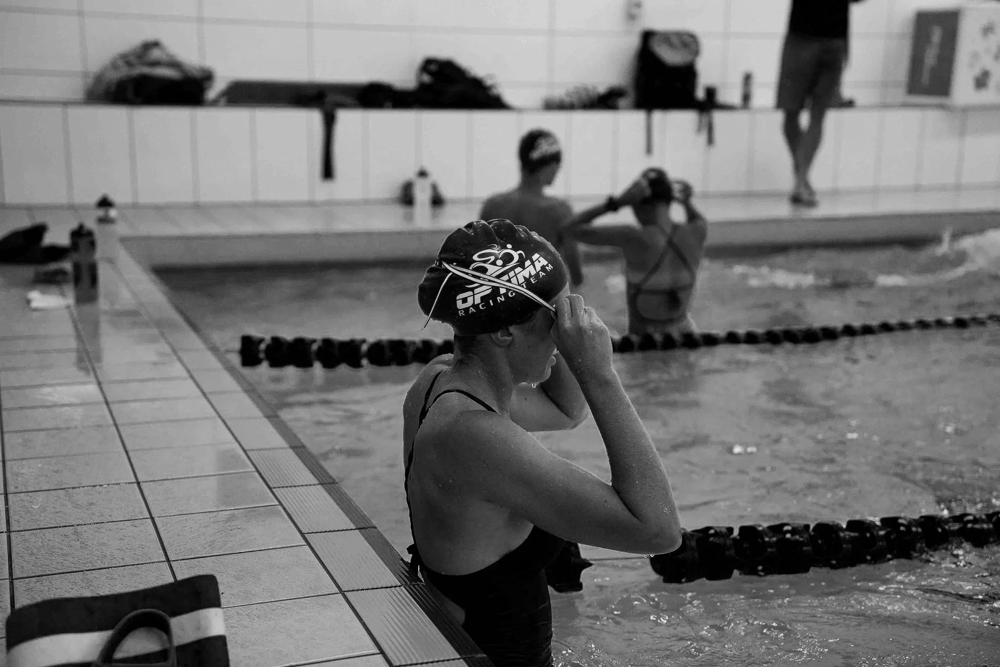 A swimmer standing at the edge of an indoor swimming pool, adjusting her swim cap, with other swimmers and a coach in the background.