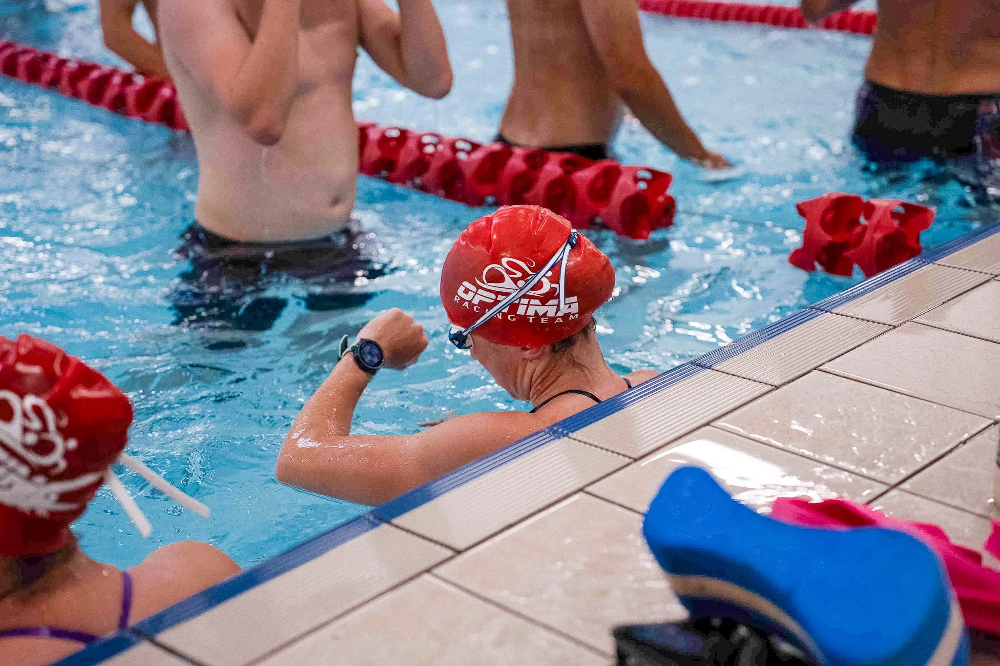 Swimmers in a swimming pool, wearing red caps with white text, during a training session or race. One swimmer is resting at the edge of the pool, while others are in the water near the lane dividers.