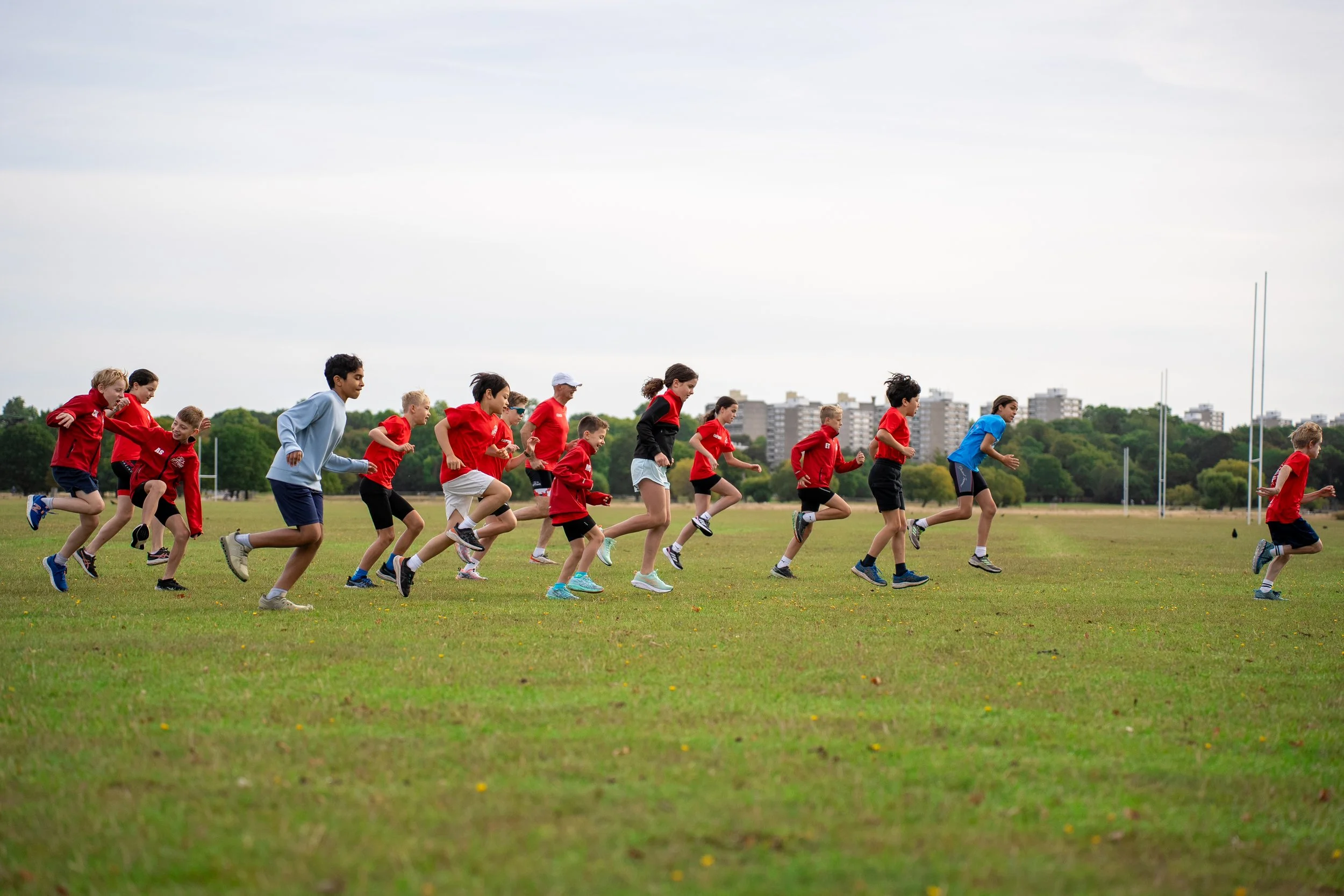 Children running on a grassy field during an outdoor sports practice, with a cityscape and trees in the background.