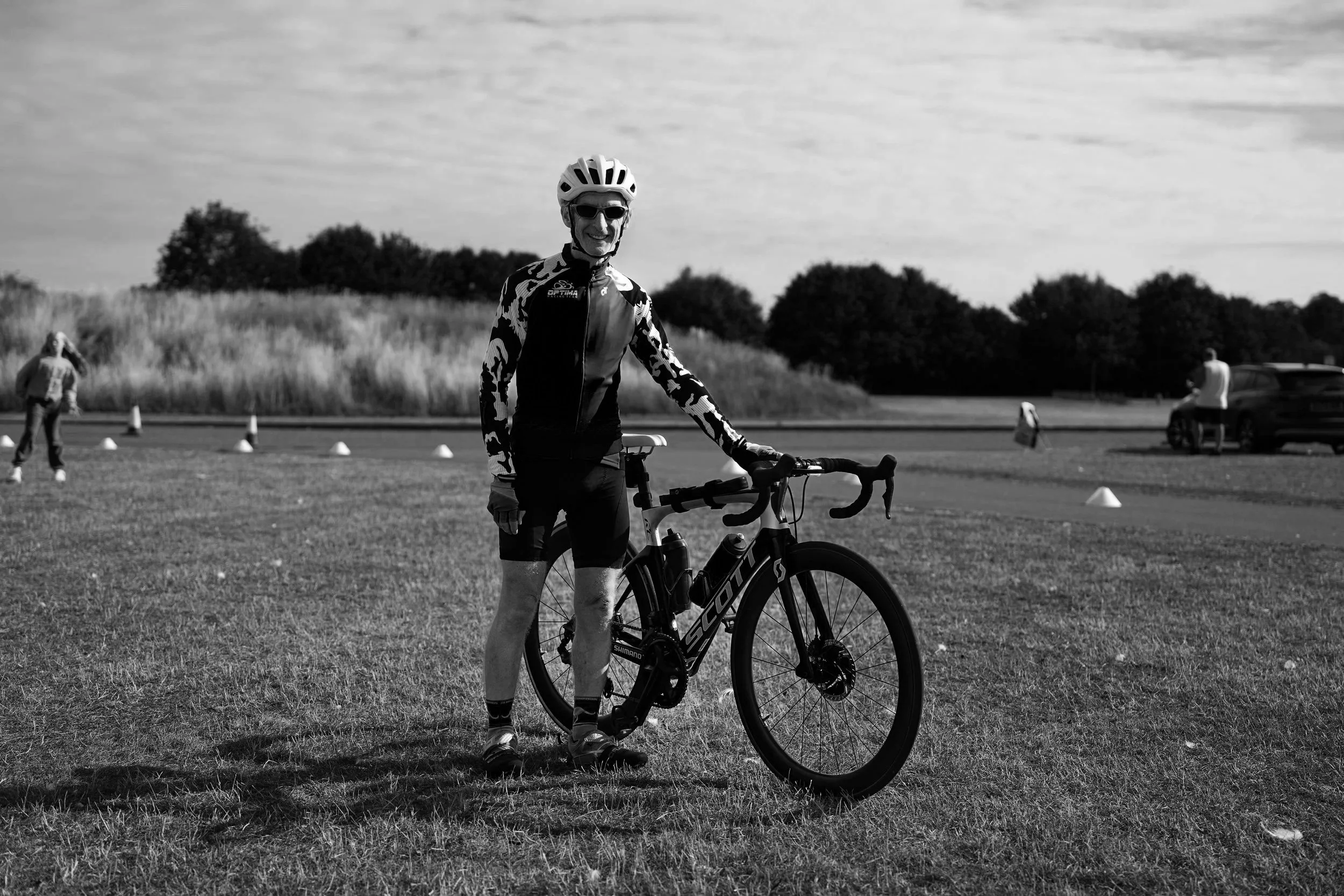 A man in cycling gear with a helmet and sunglasses standing outdoors with a bicycle in a grassy field, with other people and cars in the background.