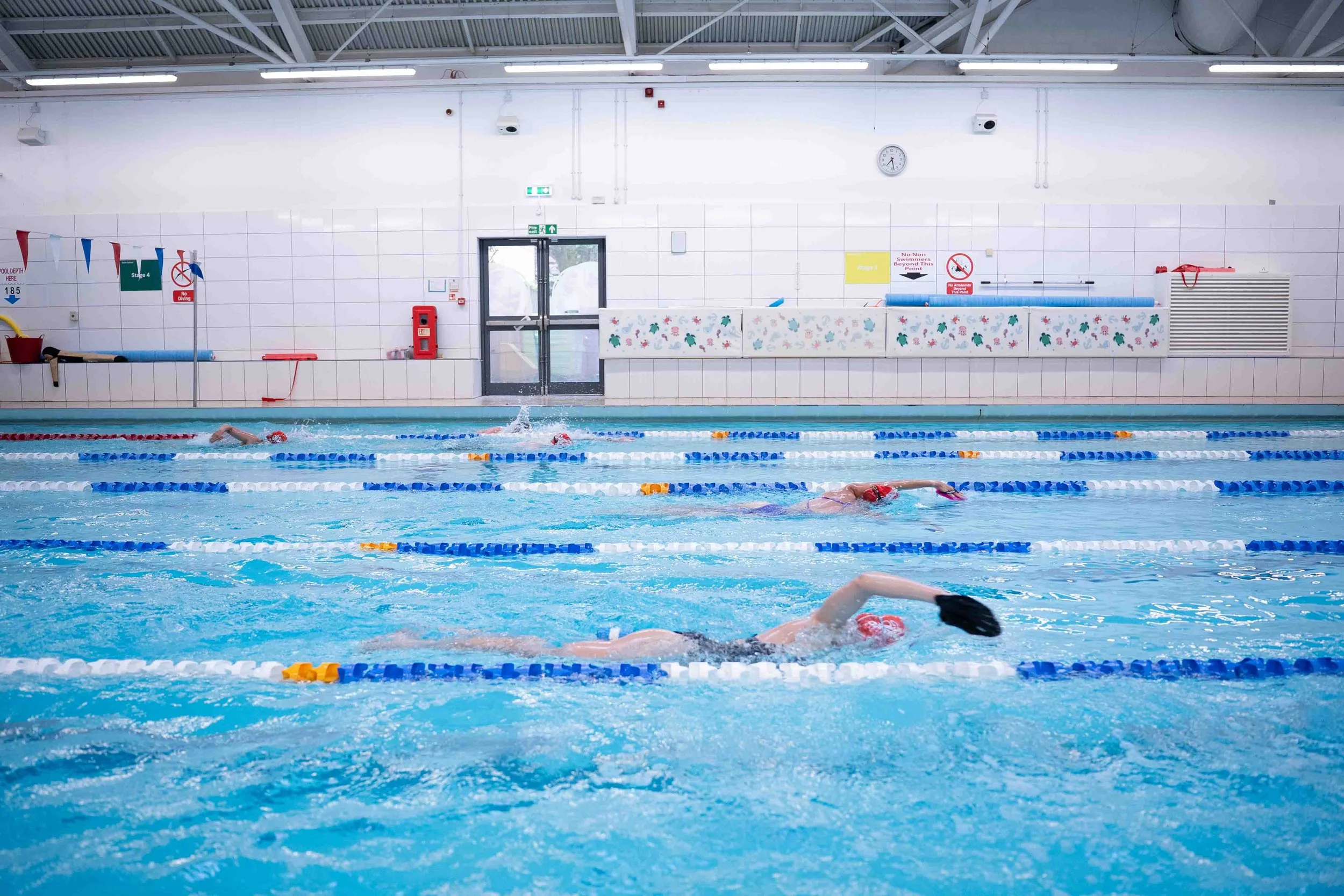 Indoor swimming pool with several children swimming laps, wearing red swim caps and goggles, under bright lighting with white tiled walls and safety signs.