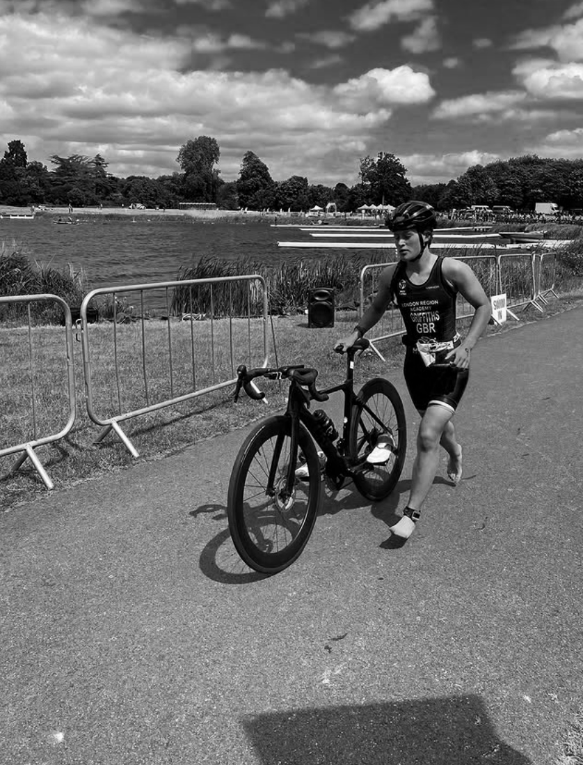 A female athlete walking along a pathway holding a black bicycle, near a lake with boats, trees, and a cloudy sky in the background.