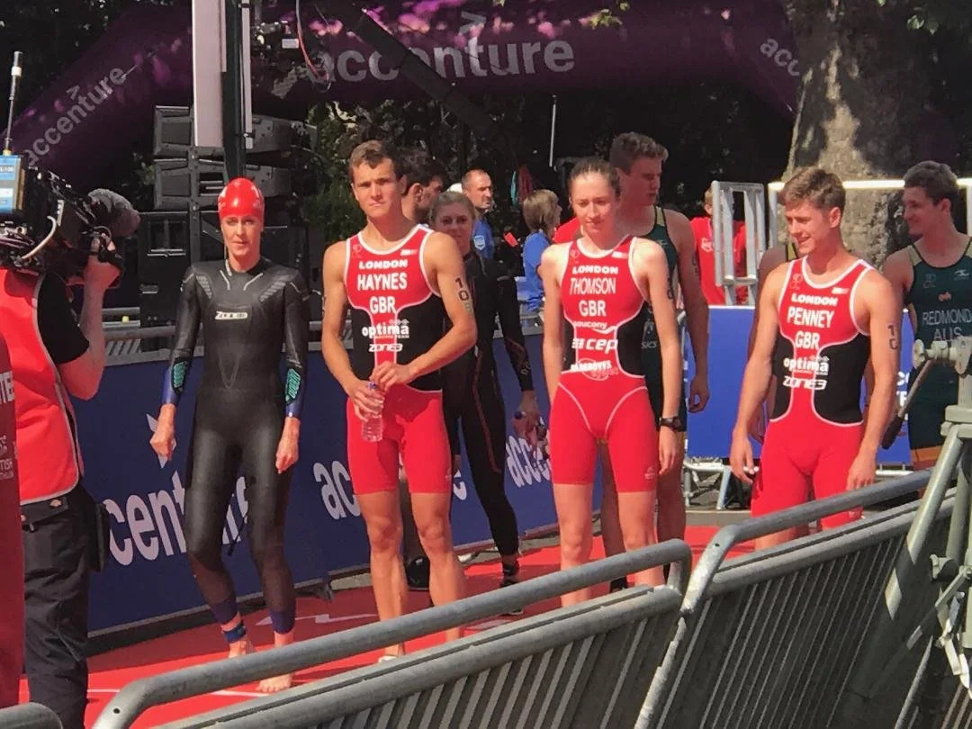 Four athletes in red triathlon suits standing on a red carpet, one woman in a black wetsuit with a red swim cap, and camera crews filming them at an outdoor event.